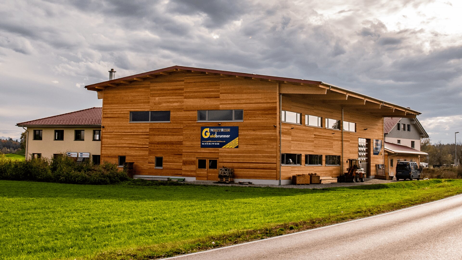 Ein großes hölzernes Geschäftsgebäude mit einem Schild mit der Aufschrift „G. Gruberbauer“ steht neben einer Landstraße, umgeben von grünem Gras unter einem bewölkten Himmel.