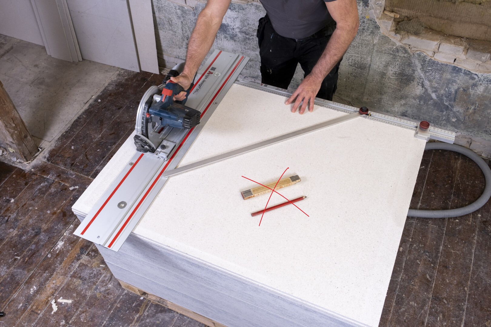 A person uses a circular saw with a guide rail to cut a large white board. A red X marks two wooden rulers on top of the board, indicating not to use them for cutting guidance.