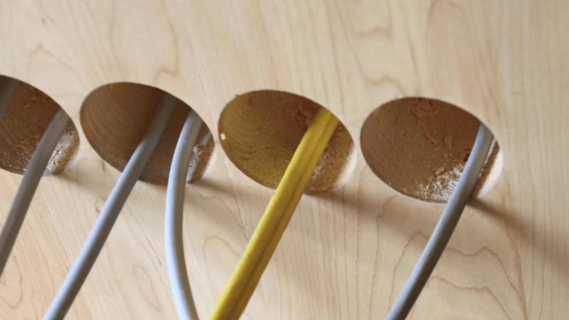 Vier Kabel – drei graue und ein gelbes – verlaufen durch runde Löcher, die in eine helle Holzoberfläche gebohrt sind. An den Rändern der Löcher sind Holzspäne sichtbar.