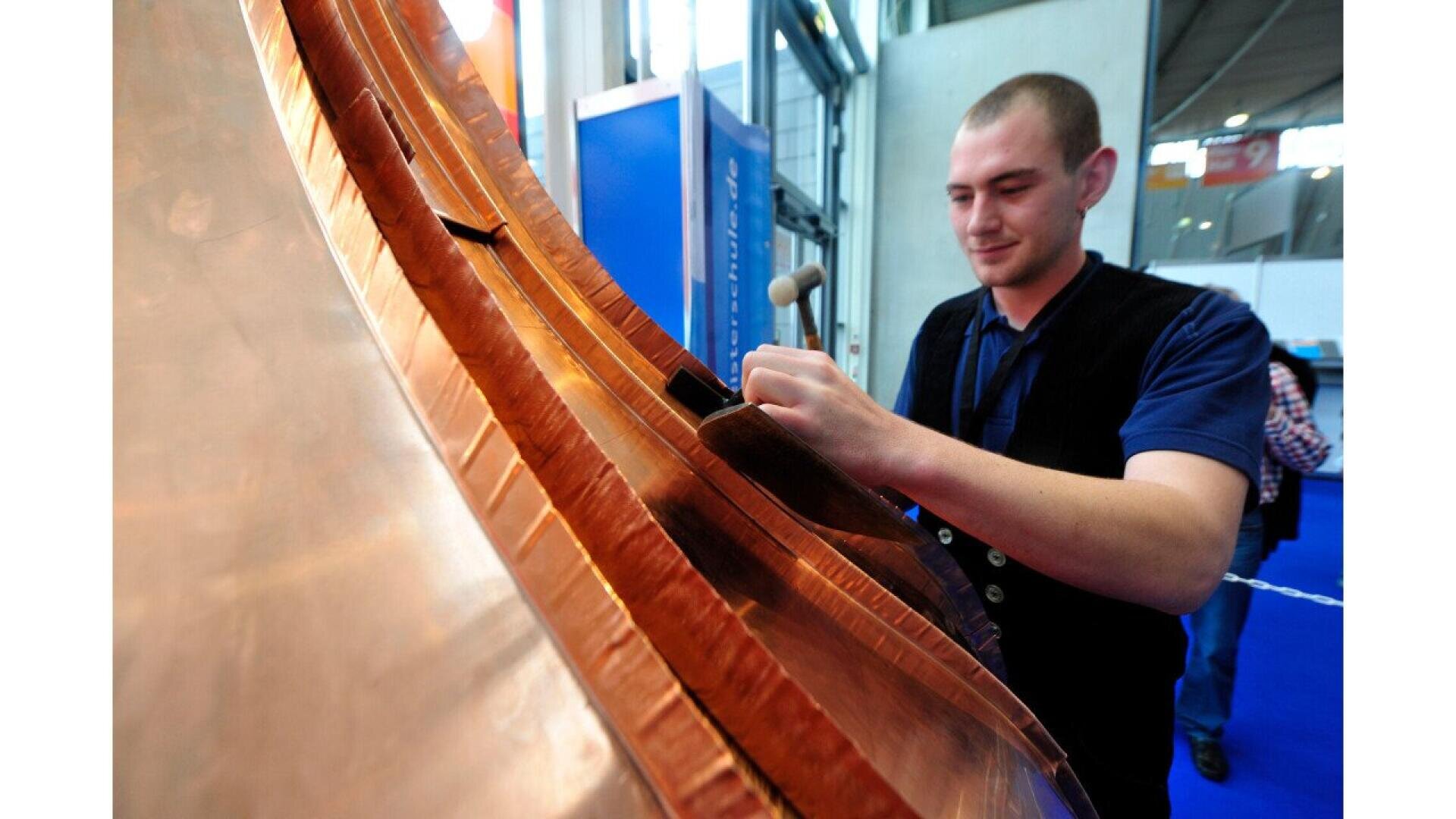 A man uses a small hammer and tool to work on a large, curved copper surface at an indoor event or exhibition, focusing intently on his task.