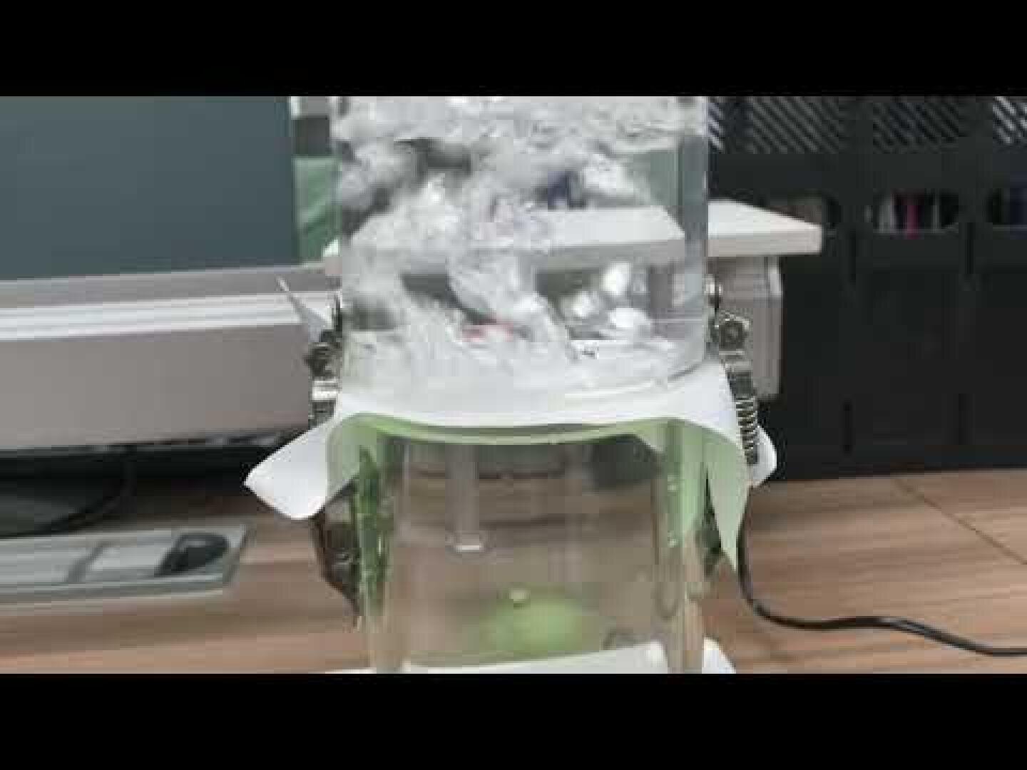 Nahaufnahme einer Laborausrüstung mit stark sprudelndem Wasser in einem durchsichtigen Behälter, der auf einem Tisch mit Computern und anderen Geräten im Hintergrund steht.