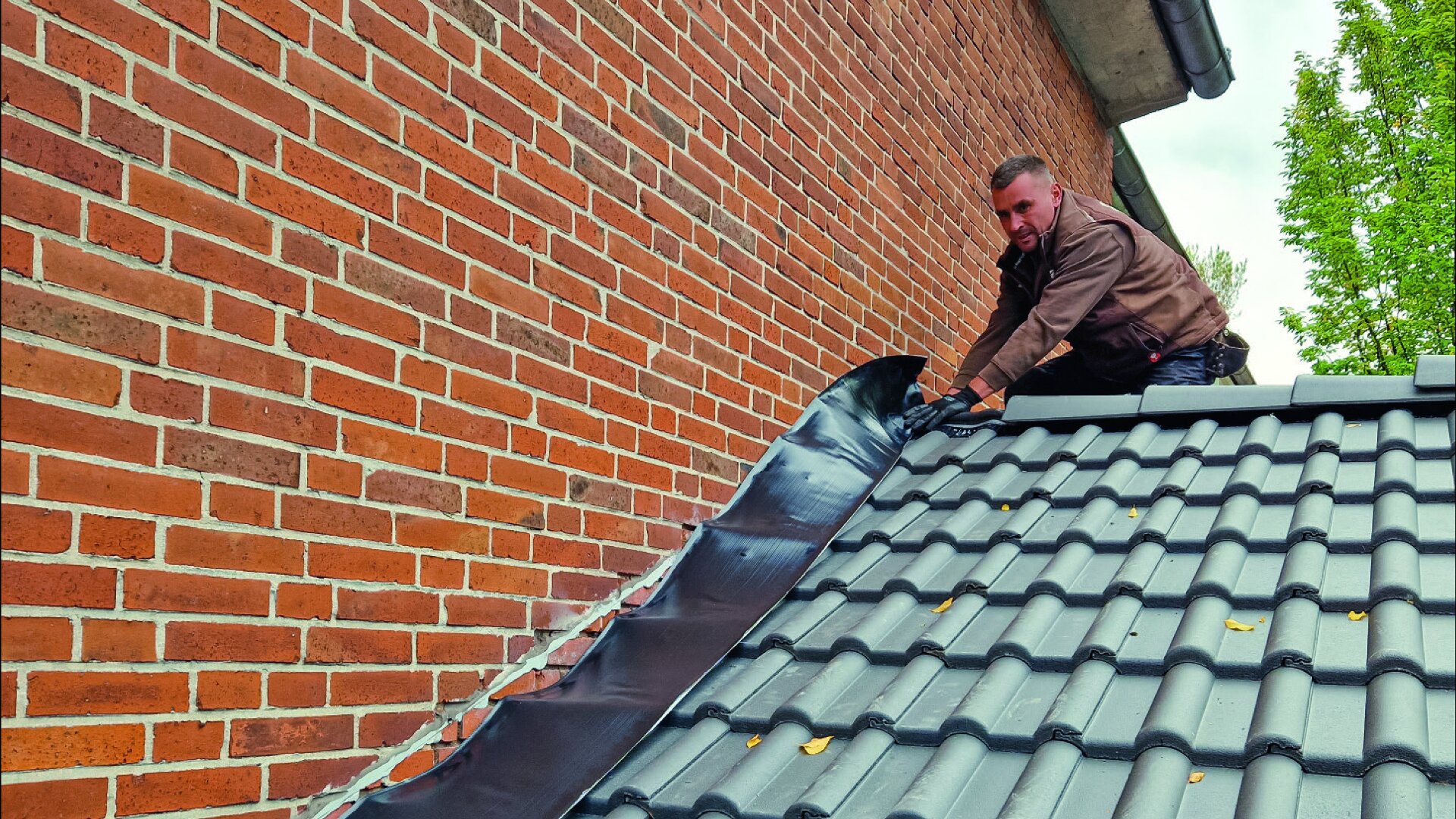Ein Mann bringt schwarze Dichtungsbahnen an der Stelle an, an der ein Ziegeldach auf eine Ziegelwand trifft, um eine wasserdichte Abdichtung zu gewährleisten. Er kniet auf dem Dach und arbeitet mit Werkzeug. Im Hintergrund sind Bäume zu sehen.
