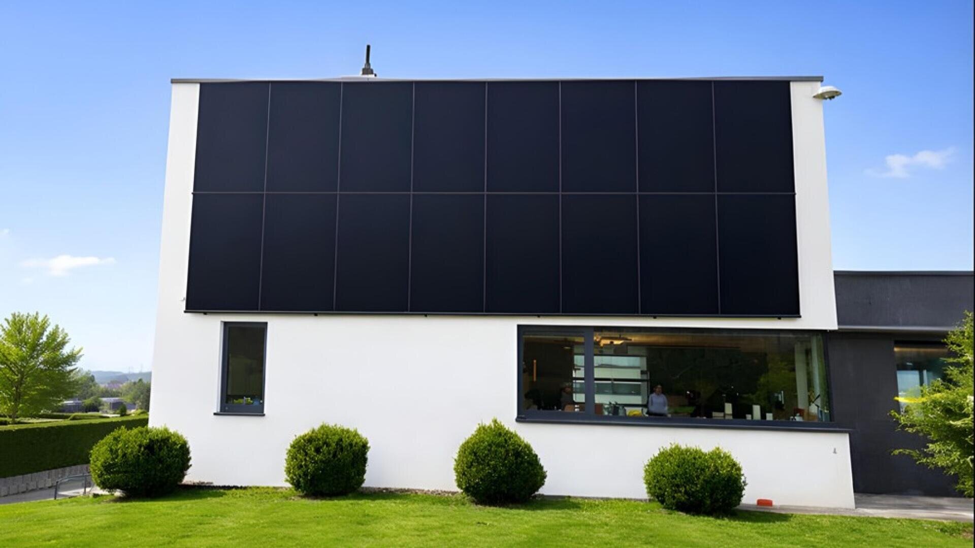 Modernes weißes Haus mit großen schwarzen Sonnenkollektoren an der oberen Hälfte der Außenwand, darunter Fenster, grüner Rasen, beschnittene Büsche und klarer blauer Himmel im Hintergrund.