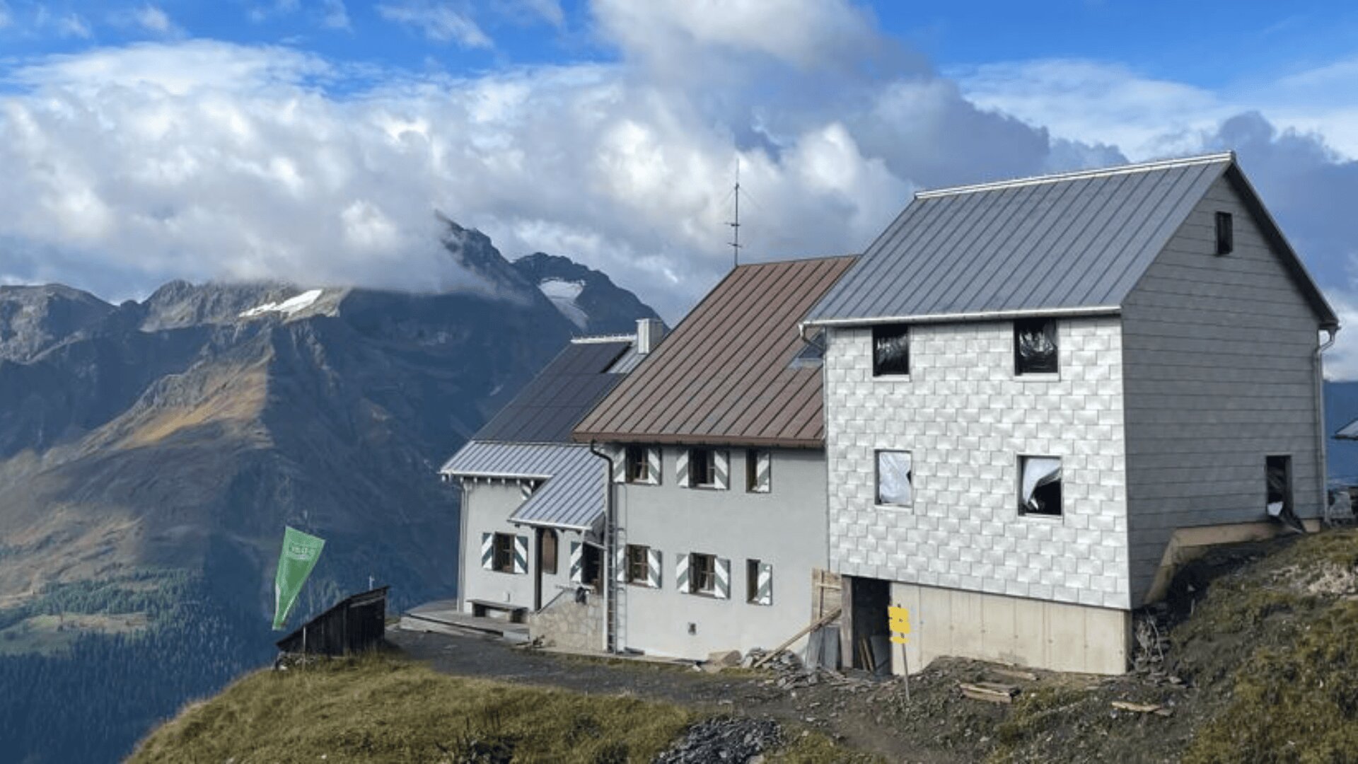 Eine Berghütte mit Metalldach und weißer Außenverkleidung steht an einem grasbewachsenen Hang mit dramatischen Alpengipfeln und bewölktem Himmel im Hintergrund. In der Nähe des Gebäudes sind eine grüne Flagge und ein Wegweiser zu sehen.