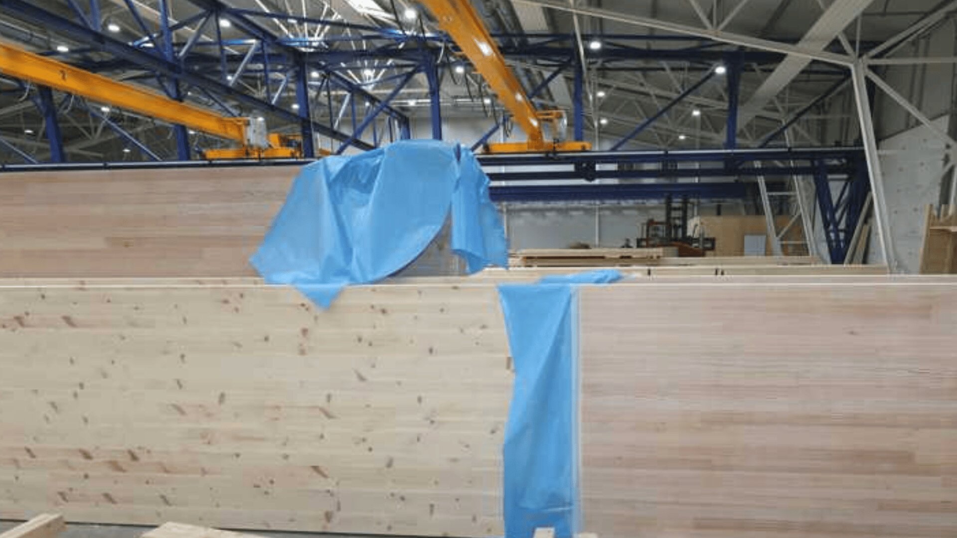 Große Holzpaneele in einer Fabrik, teilweise mit blauen Kunststofffolien verkleidet. Im Hintergrund sind Brückenkräne und Metallträger zu sehen, die eine industrielle Umgebung suggerieren.