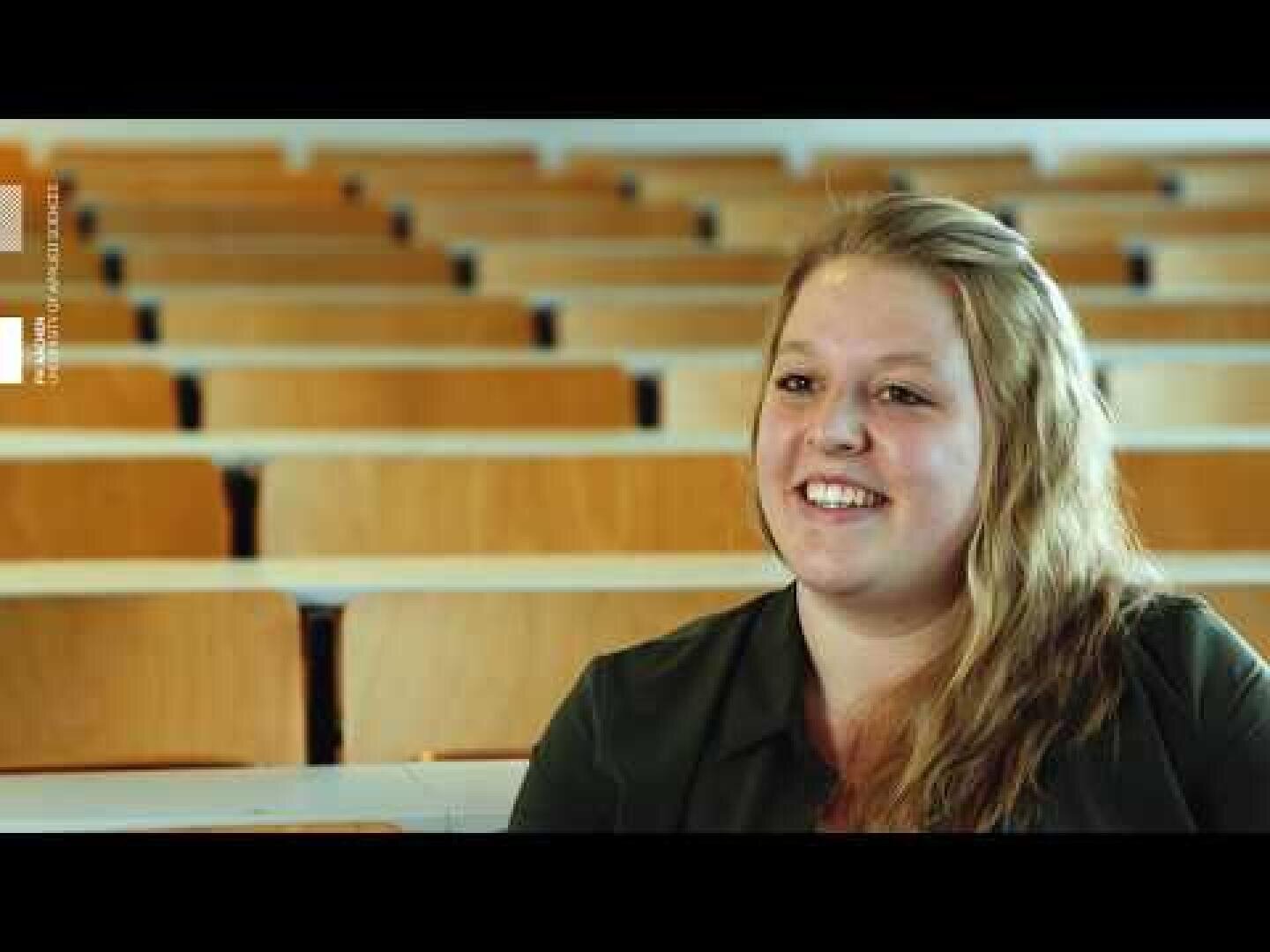 Eine junge Frau mit langen blonden Haaren sitzt lächelnd in einem leeren Hörsaal mit Reihen von Holzstühlen hinter ihr.