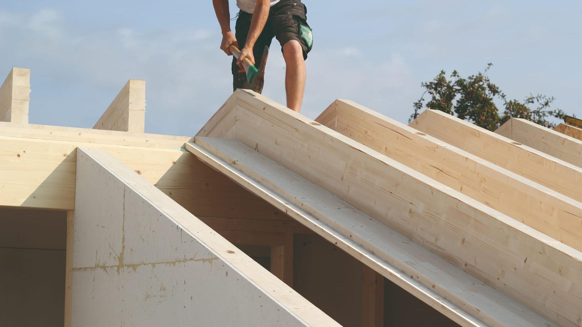 An einem sonnigen Tag arbeitet eine Person mit einem Hammer am Holzrahmen eines im Bau befindlichen Hausdachs.