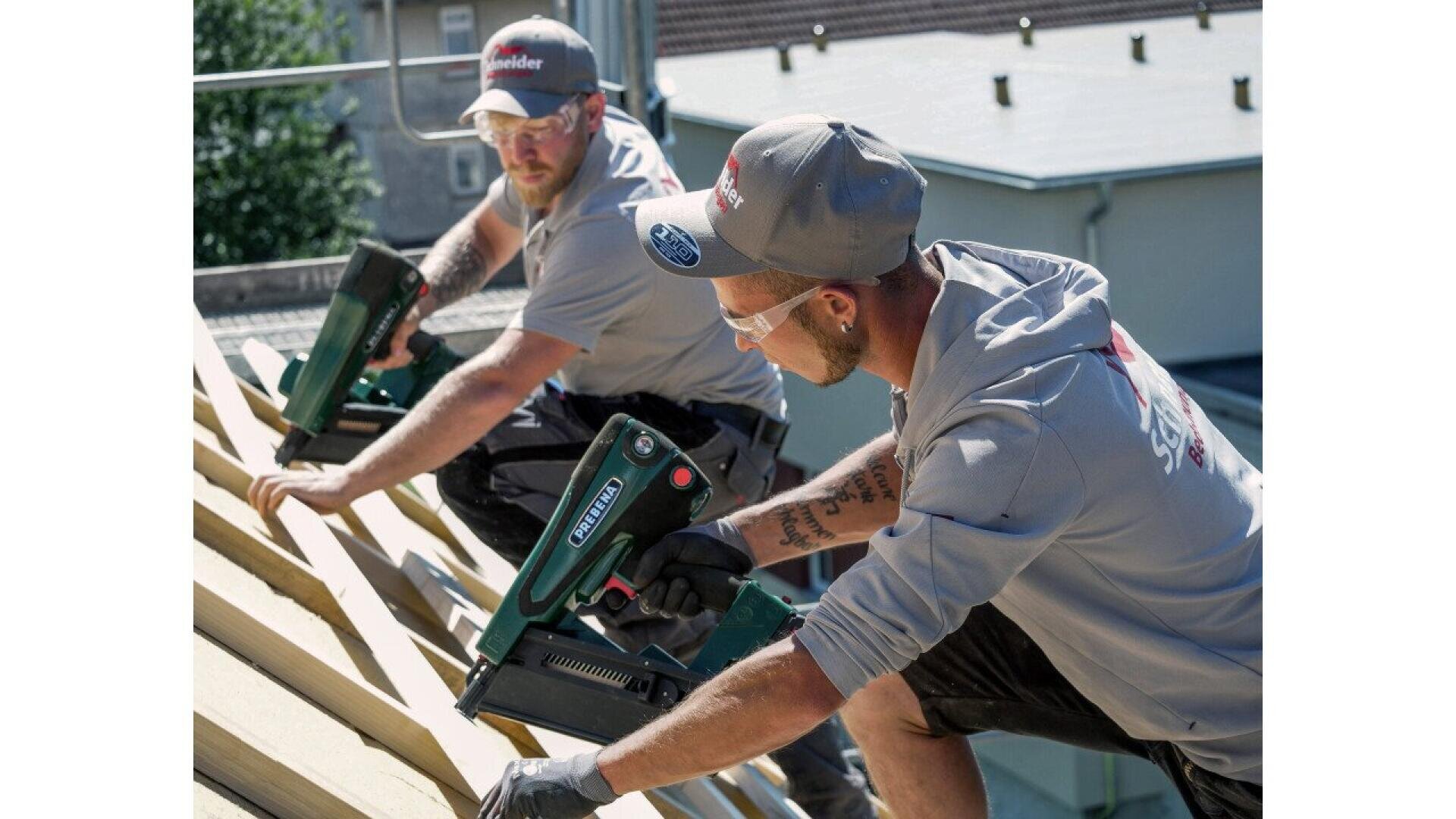 Two construction workers wearing safety glasses and caps use nail guns to attach wooden boards on a sloped roof structure, working outdoors on a sunny day.