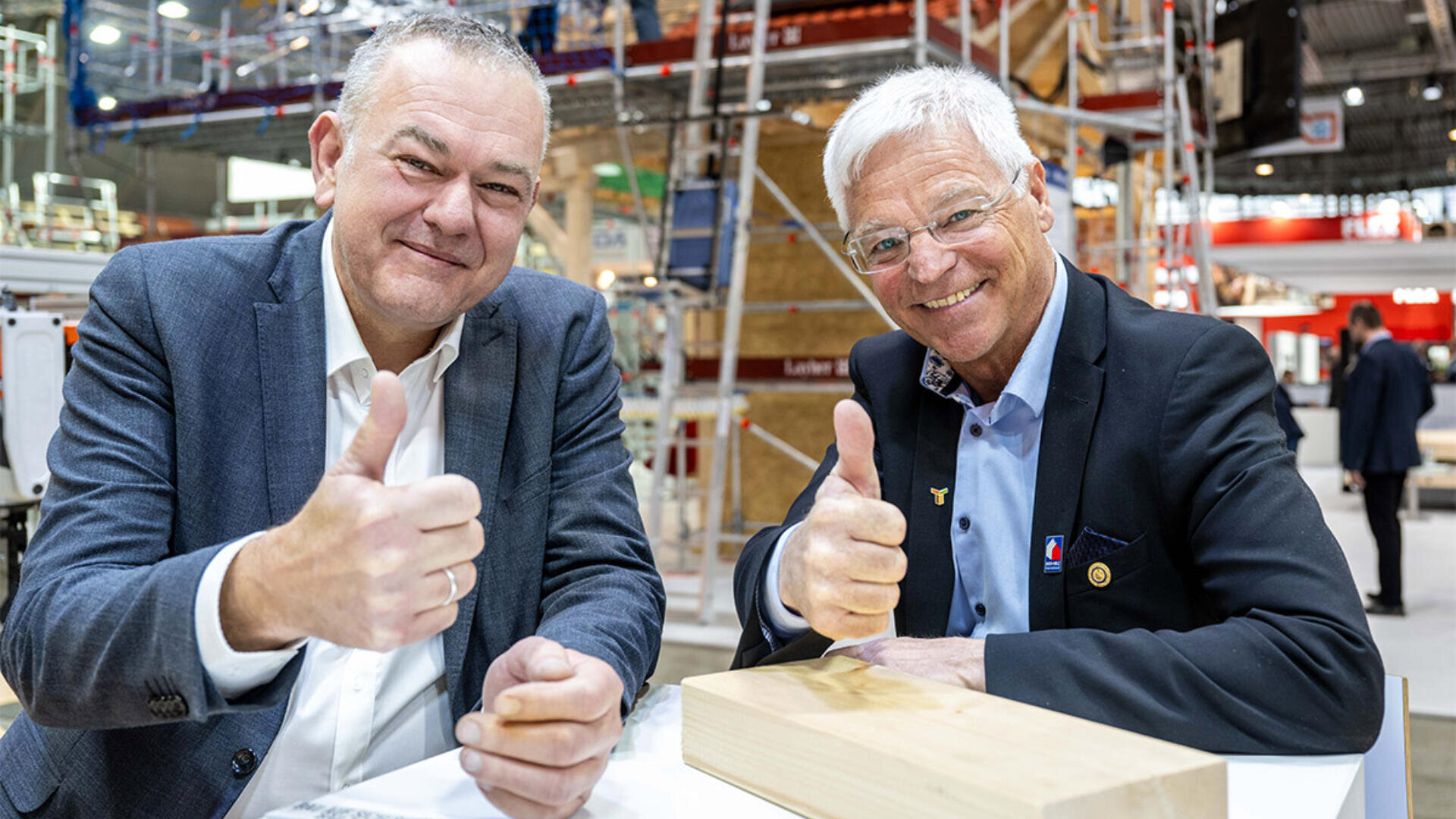 Zwei ältere Männer in Anzügen sitzen lächelnd an einem Tisch und strecken den Daumen in die Kamera. Sie scheinen sich auf einer Veranstaltung oder Messe zu befinden. Im Hintergrund sind Baumaterialien und -geräte zu sehen.