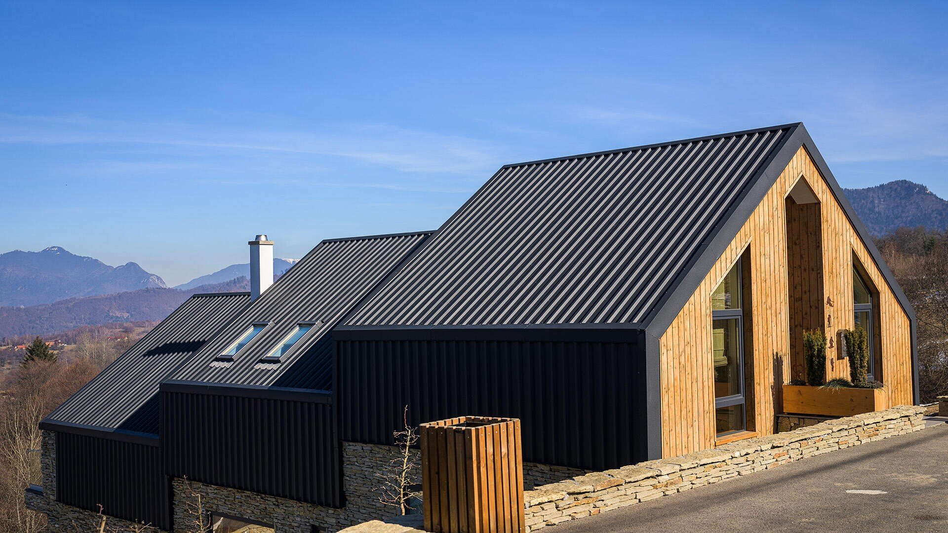 A modern house with black metal roofs and wooden walls stands on a stone terrace in a mountainous landscape under a blue sky.