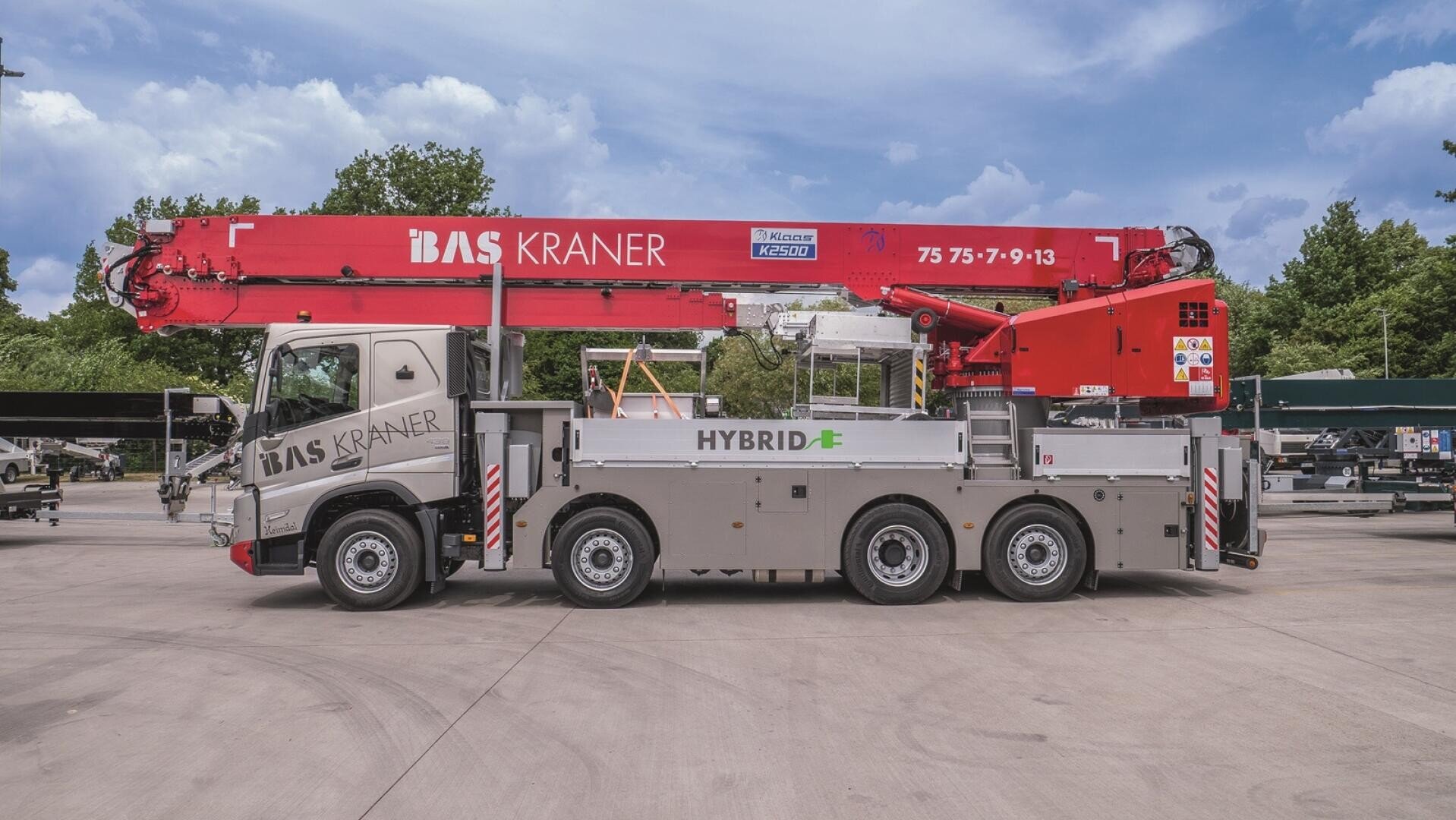 A large hybrid mobile crane truck with a red crane arm and BAS KRANER lettering is parked on a concrete site, with trees and other vehicles in the background.