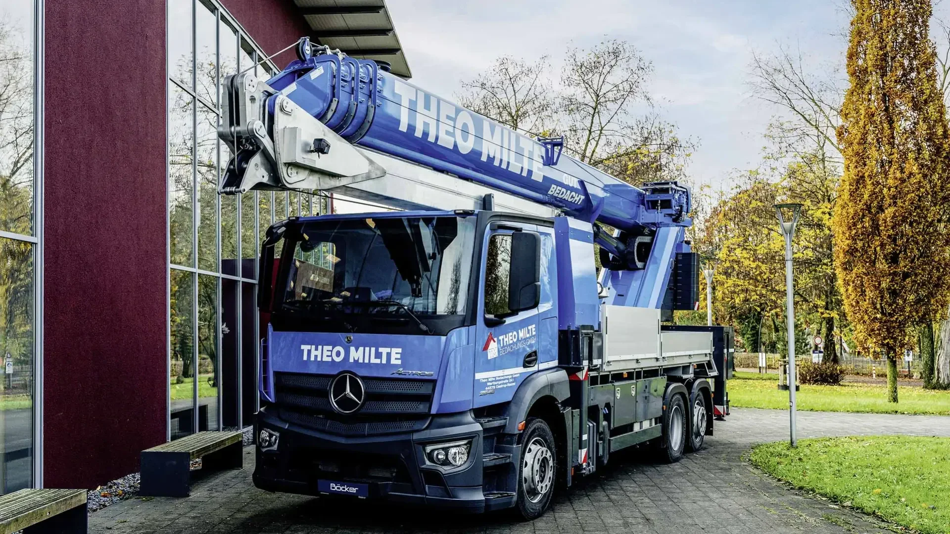 Ein blau-weißer Autokran mit der Aufschrift „THEO MILTE“ auf dem Ausleger steht auf einem gepflasterten Weg in der Nähe eines modernen Glasgebäudes und Bäumen mit Herbstlaub.