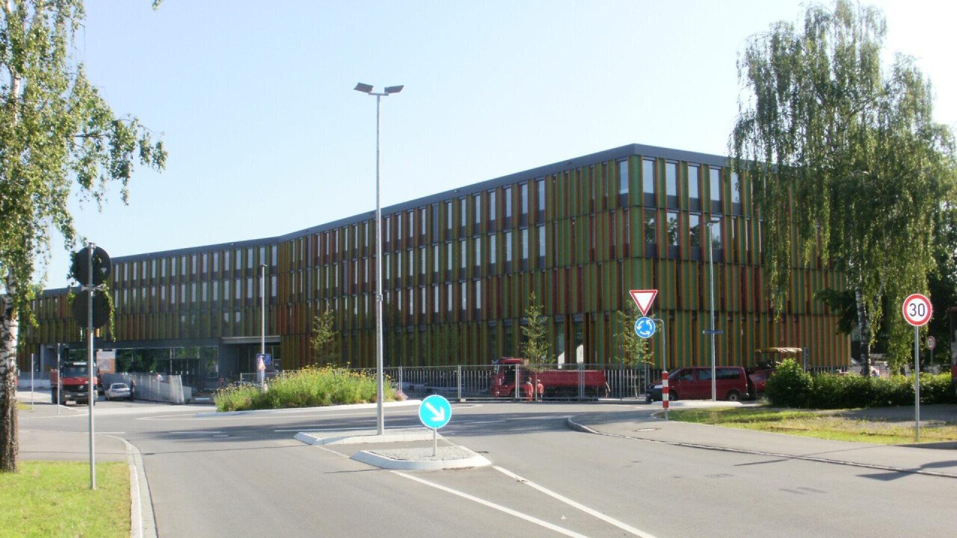 A modern, three-story building with vertical colored panels stands by a street. There are traffic signs, a roundabout, and some trees and parked vehicles near the building. The sky is clear and sunny.
