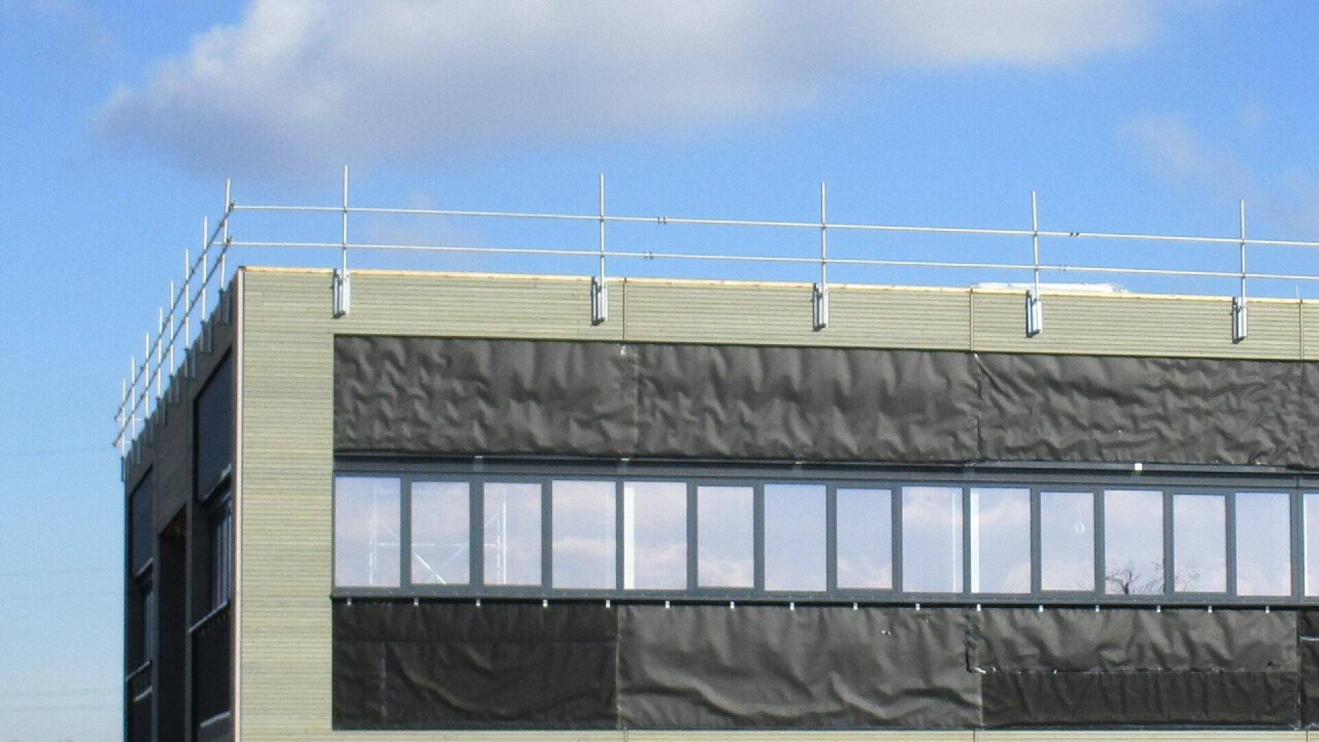 The top floor of a modern building with large windows and a temporary safety railing along the roof edge, against a blue sky with clouds.