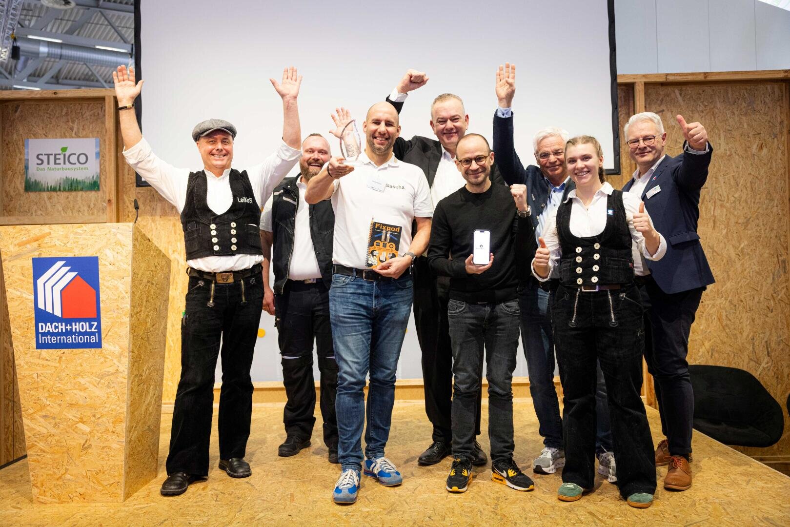 Eine Gruppe von acht lächelnden Menschen steht auf einer Bühne, einige mit feierlich erhobenen Armen. Zwei Personen in der ersten Reihe halten Auszeichnungen. Ein DACH+HOLZ International-Schild ist auf dem Podium zu sehen.