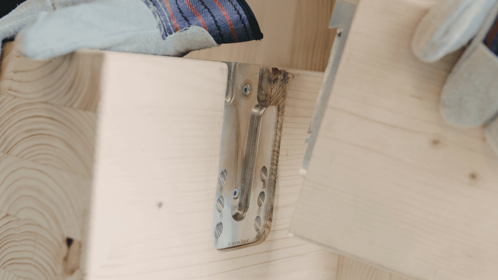 A close-up of a metal bracket securing two pieces of light-colored wood, with a gloved hand holding one piece in place.