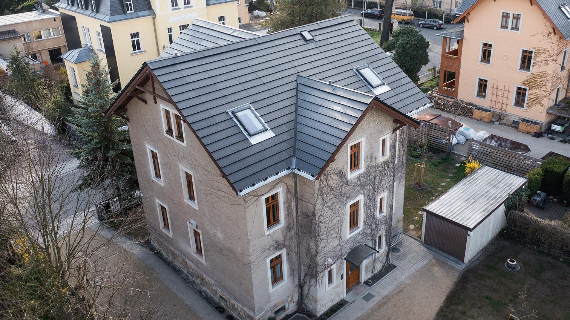 Luftaufnahme eines dreistöckigen beigen Hauses mit grauem Schrägdach, mehreren Oberlichtern und mehreren Fenstern, umgeben von Bäumen, anderen Häusern und einer kleinen Einfahrt mit einem Schuppen in der Nähe.