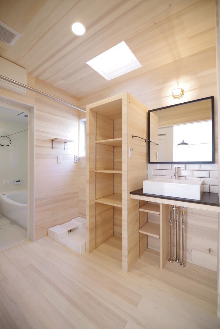 Ein modernes Badezimmer mit hellen Holzwänden und -böden, offenen Regalen, einem rechteckigen Waschbecken mit freiliegenden Rohren, einem großen Spiegel, einer weißen Fliesenrückwand und Blick in einen weißen Badewannenbereich. Ein Oberlicht erhellt den Raum.