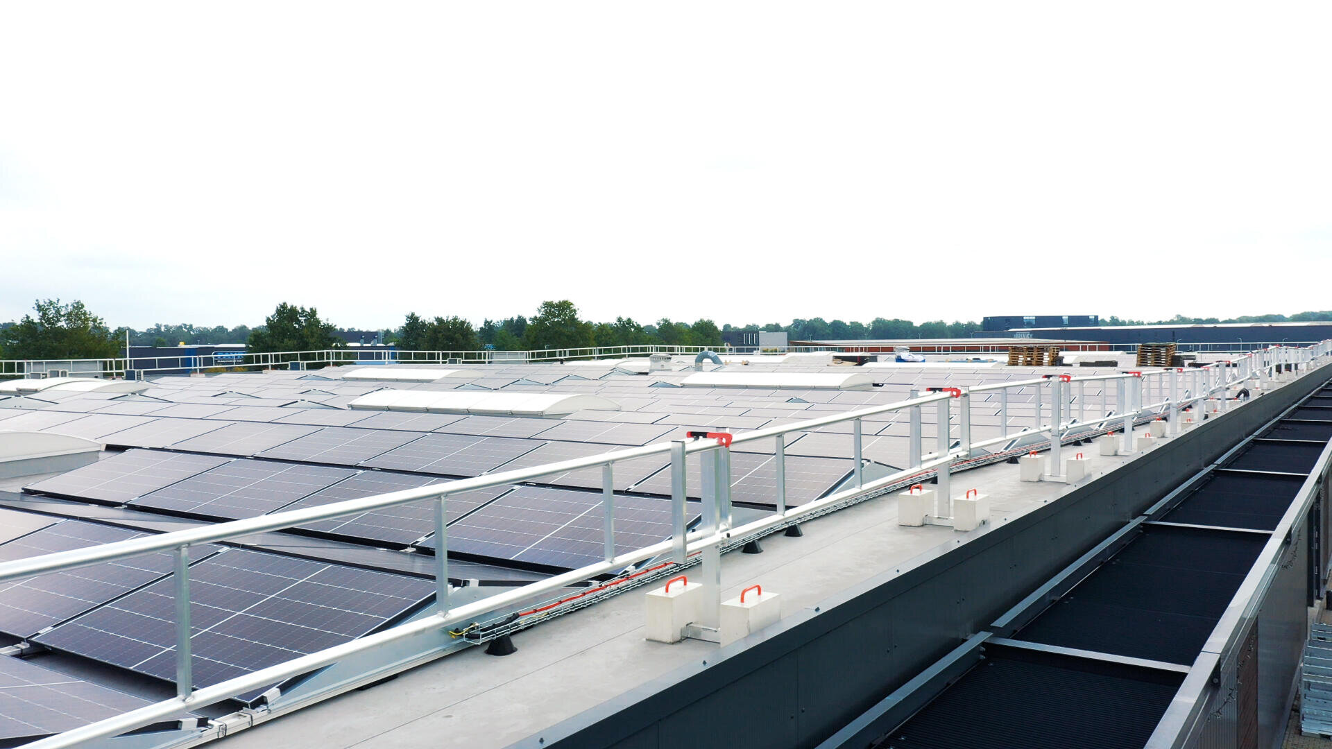 Solar cells installed on the roof of a flat building, surrounded by safety railings, with trees and a cloudy sky in the background.