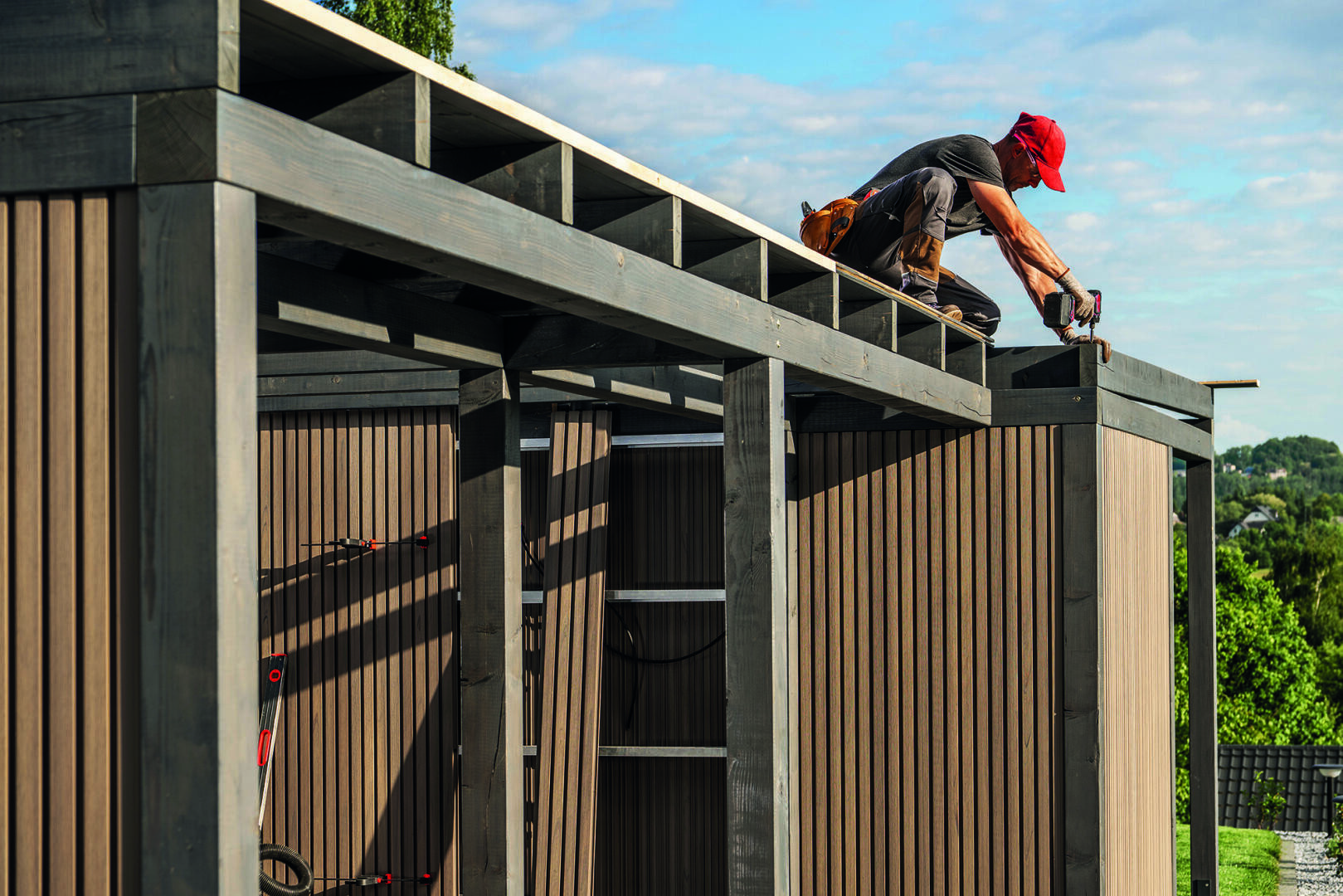 Eine Person mit einer roten Mütze kniet auf dem Dach eines teilweise errichteten Holzschuppens und benutzt ein Elektrowerkzeug. Der Schuppen steht an einem sonnigen Tag im Freien, umgeben von Grün.