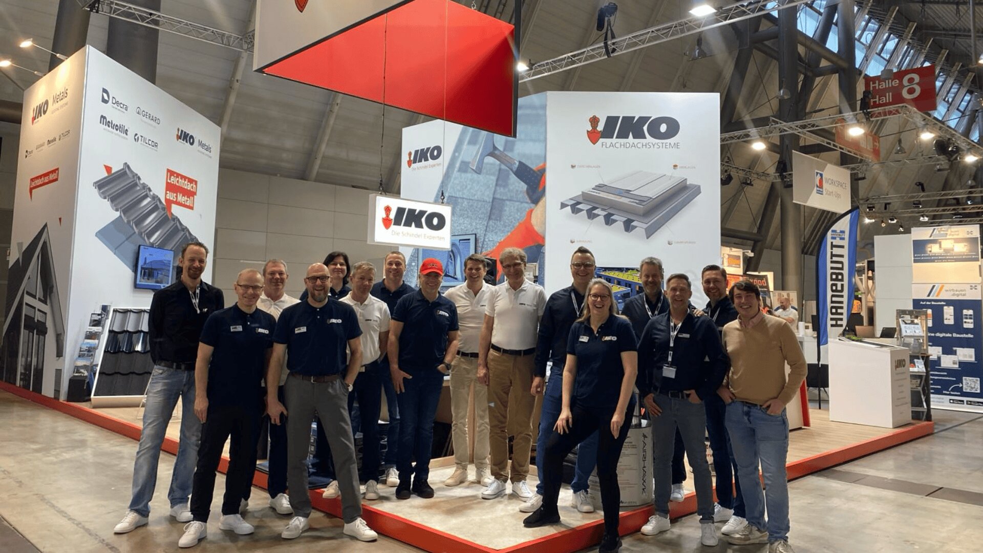 Eine Gruppe von sechzehn Personen in Freizeit- und Businesskleidung steht lächelnd vor einem Stand von IKO Roofing Systems auf einer Indoor-Messe oder Ausstellung. Im Hintergrund sind Schautafeln und Schilder zu sehen.