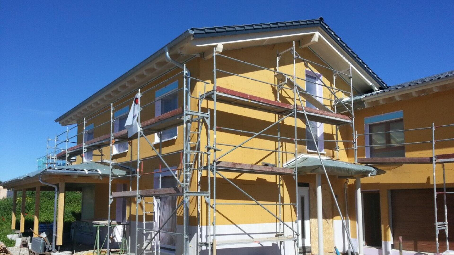A yellow house under construction, surrounded by metal scaffolding, with clear blue sky in the background. The exterior walls are unfinished, and construction materials are visible around the building.