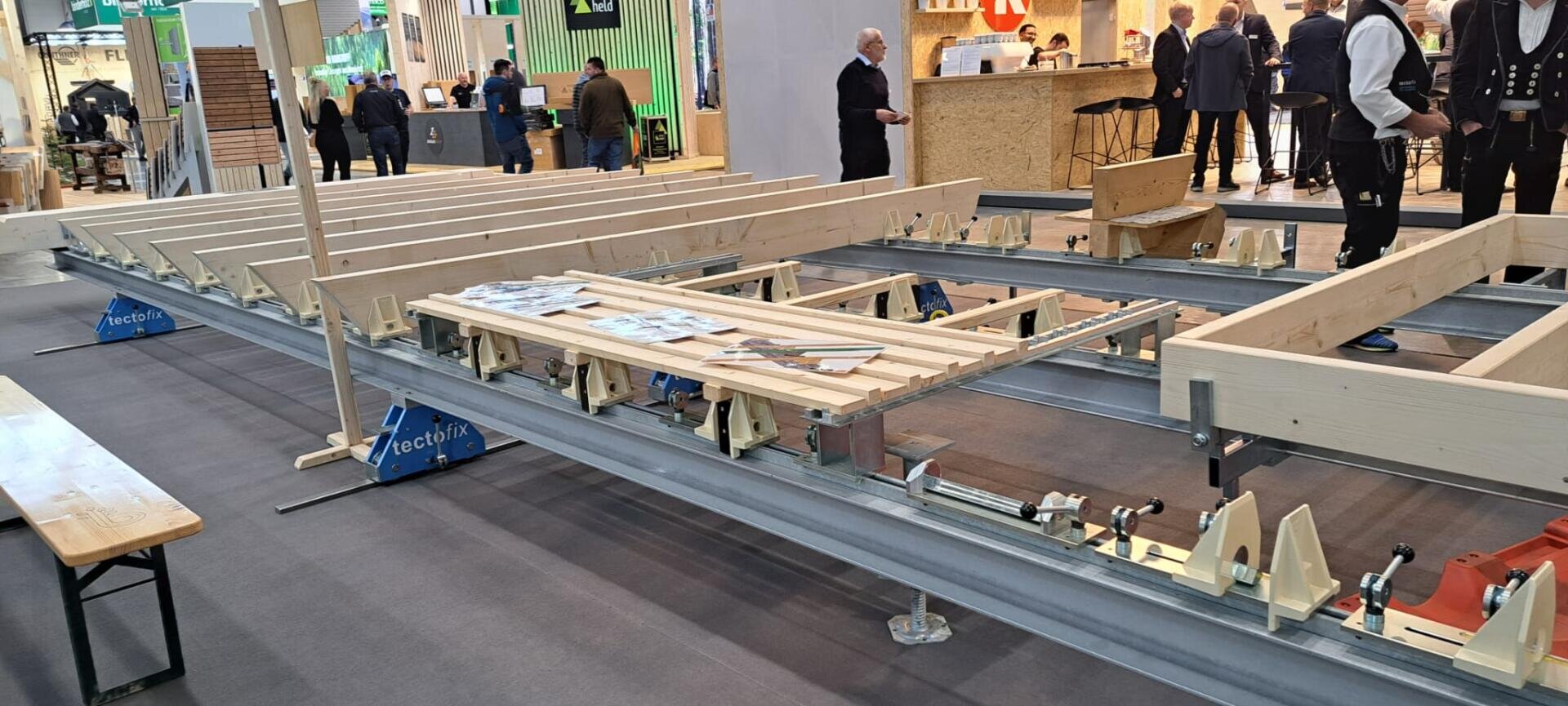 Holzbalken und -bretter sind auf einem Metallrahmen mit Klammern auf einer Indoor-Ausstellung angeordnet. Im Hintergrund stehen und gehen Menschen in der Nähe von hölzernen Ständen und Auslagen. Auf dem Holz im Vordergrund sind Flugblätter angebracht.