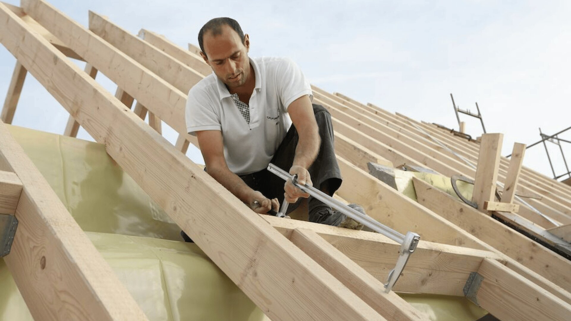 Ein Bauarbeiter im weißen Hemd befestigt mit einem Hammer Balken, während er unter klarem Himmel den Holzrahmen eines Daches baut.