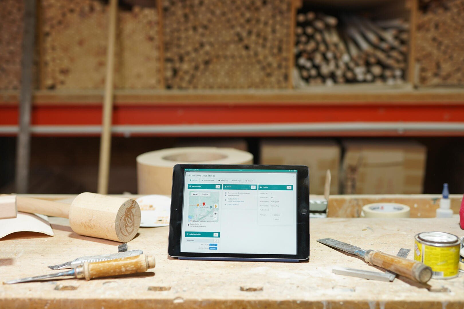 Ein Tablet mit einer Mapping-Anwendung steht auf einer Werkbank in einer Holzwerkstatt, umgeben von Holzbearbeitungswerkzeugen und Holzmaterialien, die in Regalen im Hintergrund gelagert sind.