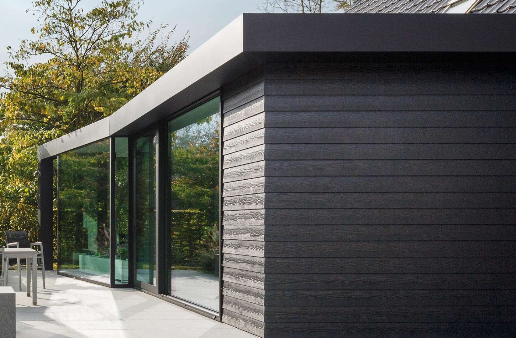 Ein moderner Erweiterungsbau mit dunkler horizontaler Holzverkleidung, großen, raumhohen Glastüren und einem schlanken Flachdach, umgeben von Bäumen und Gartenmöbeln auf einer Terrasse.