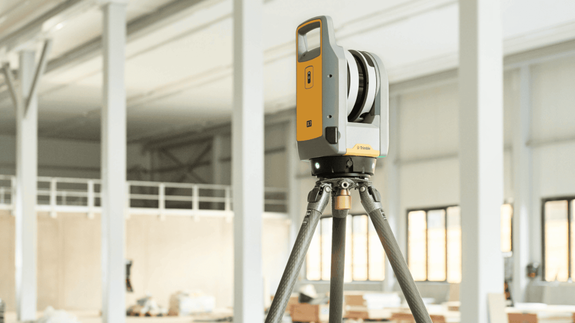A yellow and gray 3D laser scanner is mounted on a tripod inside a bright, modern industrial building with large windows and white structural beams.