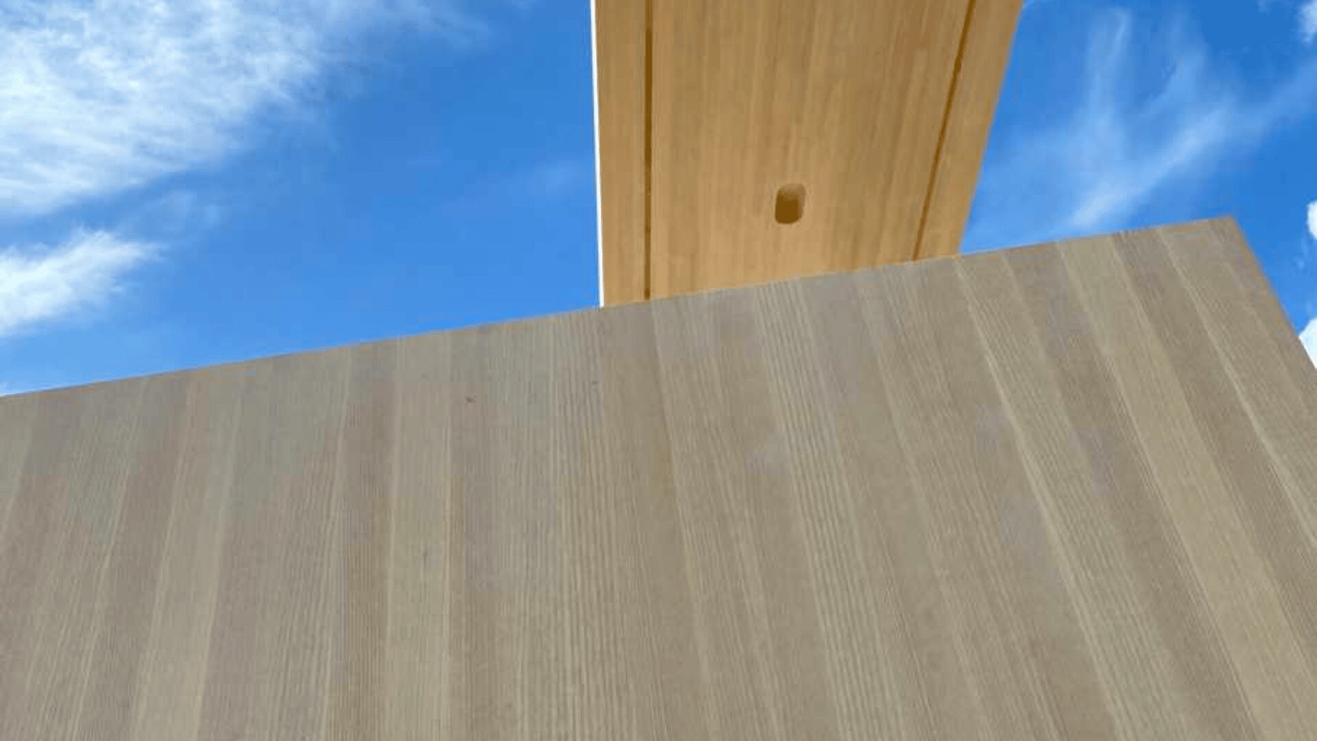 Eine Nahaufnahme großer Holzbalken und einer Wand vor einem strahlend blauen Himmel mit zarten Wolken. Das Bild hebt die Textur und Maserung der Holzpaneele hervor.
