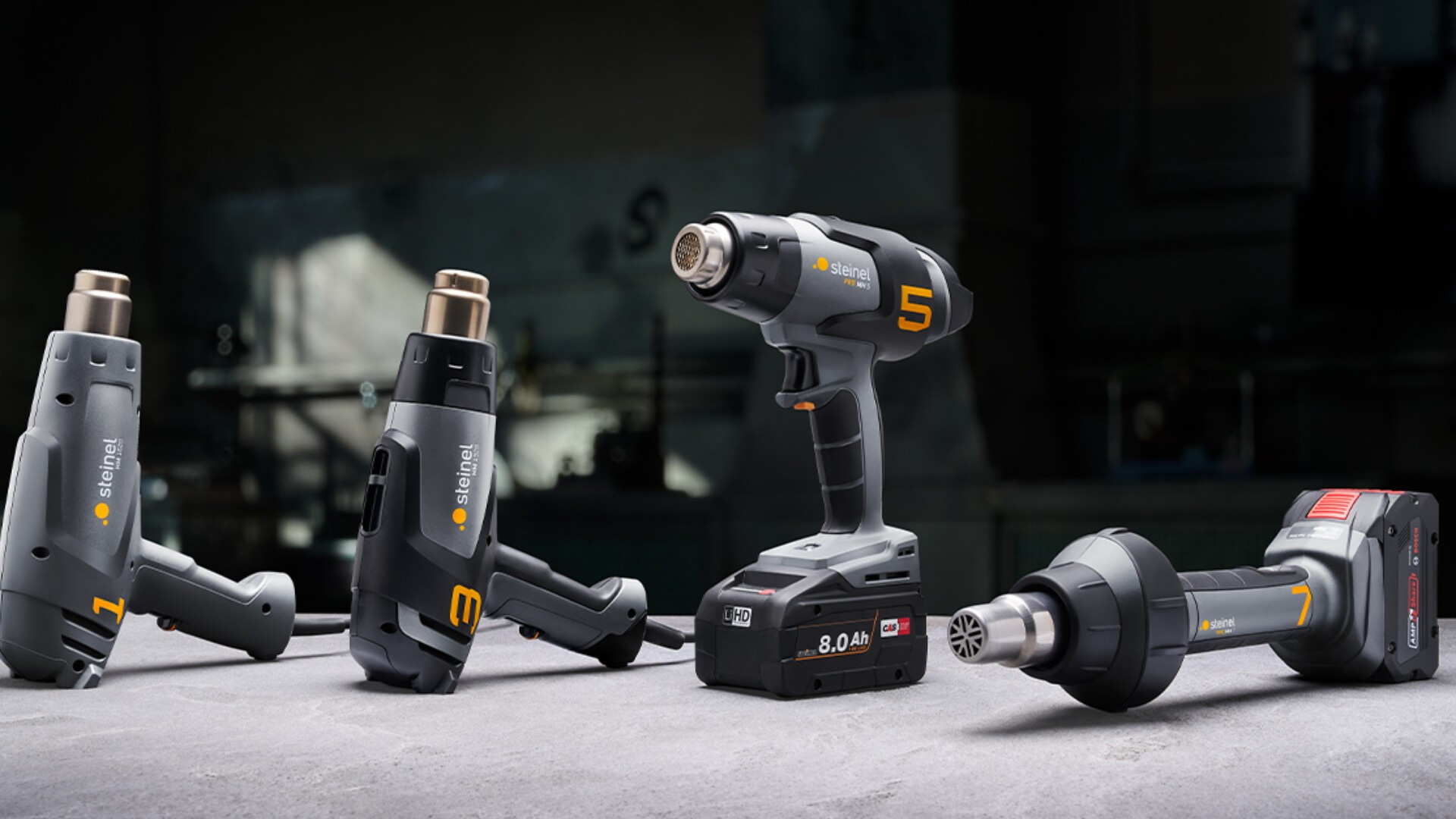 Vier verschiedene Heißluftpistolen und eine Batterie von Steinel sind auf einer grauen Oberfläche in einer Werkstattumgebung angeordnet, wobei auf jedem Werkzeug vor einem unscharfen industriellen Hintergrund Nummern und Markennamen zu sehen sind.