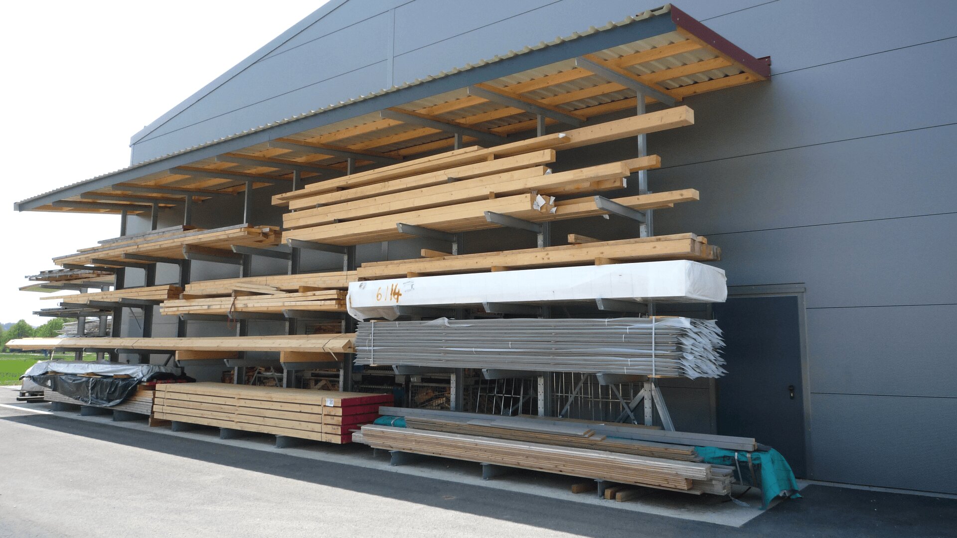 Stapel von Holzbalken, Brettern und Metallmaterialien sind auf Regalen vor einem grauen Industriegebäude unter einem überdachten Vordach geordnet. Darunter lagern auf Paletten verschiedene Baumaterialien.
