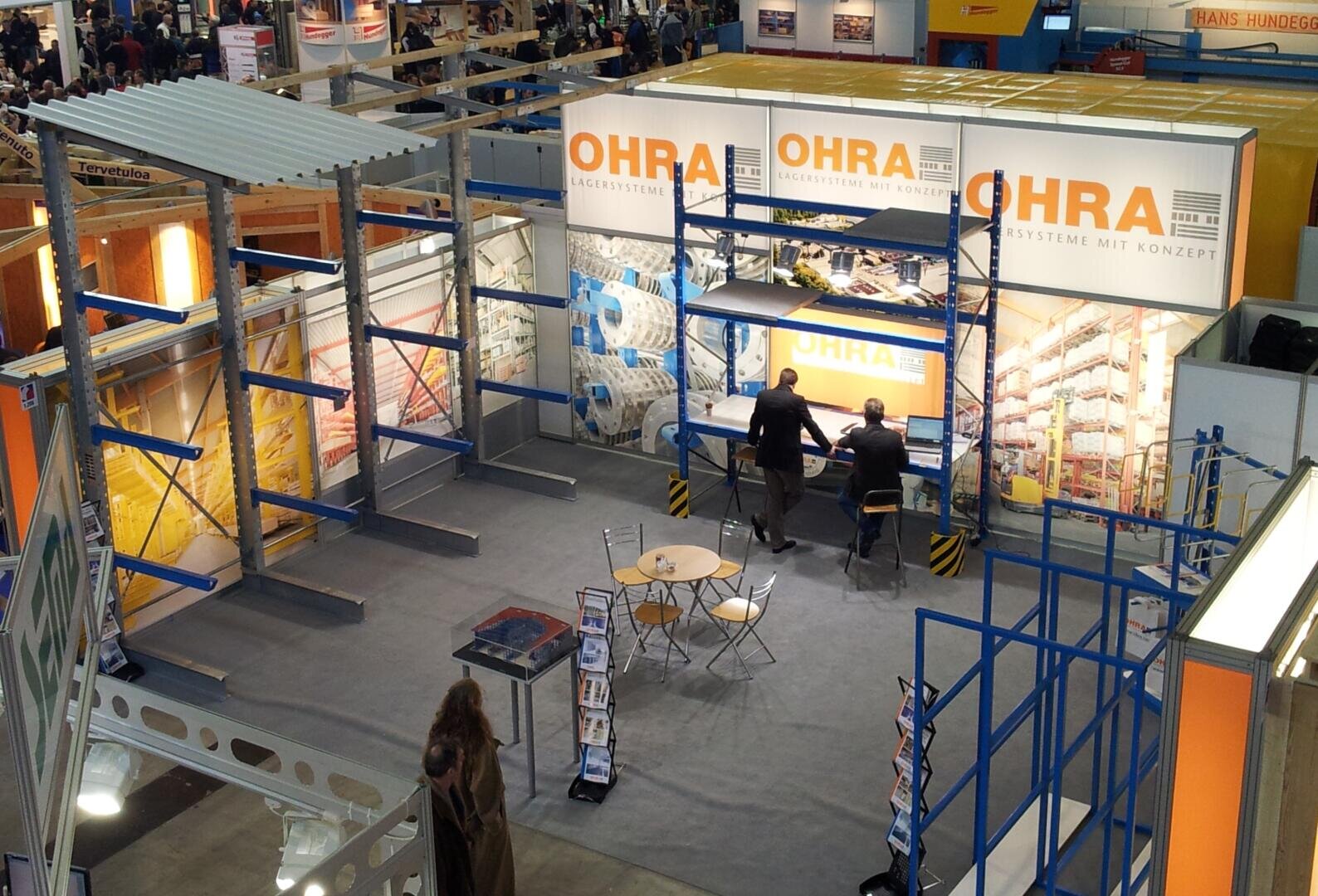 Messestand für OHRA mit Regalen, orangefarbener Beschilderung, zwei Personen an einer Theke, leeren Stühlen und einem Tisch sowie Broschüren auf einem Ständer in einer Ausstellungshalle.