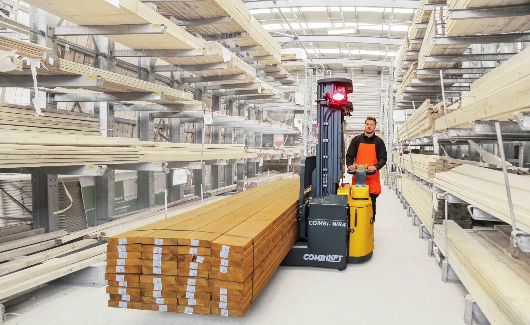 A worker in an orange safety vest operates a forklift, transporting a stack of long wooden boards through an indoor lumber warehouse with shelves full of wood panels.