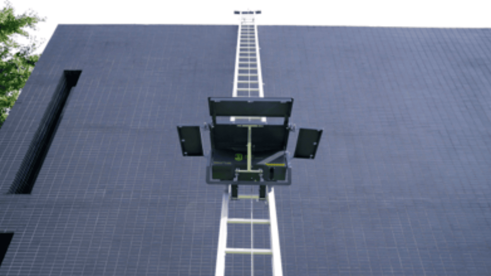 A tall, narrow metal ladder extends vertically up the side of a dark, tiled building. At the center, a black platform or hatch is attached to the ladder. The sky above is overcast.