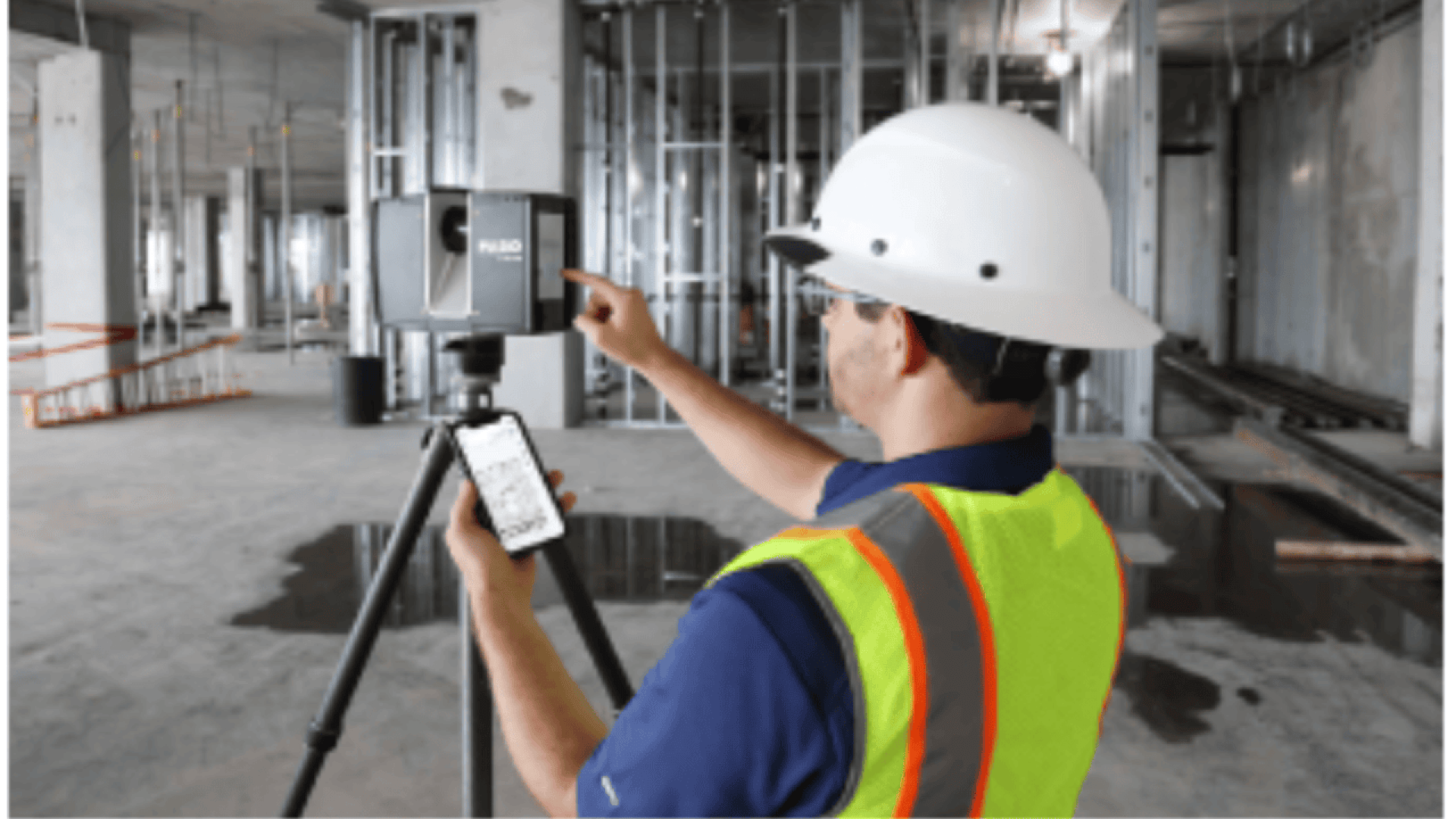 A person in a white hard hat and yellow safety vest operates a tripod-mounted laser scanner in an unfinished building, using a smartphone to control the device. Metal framing and construction materials are visible.