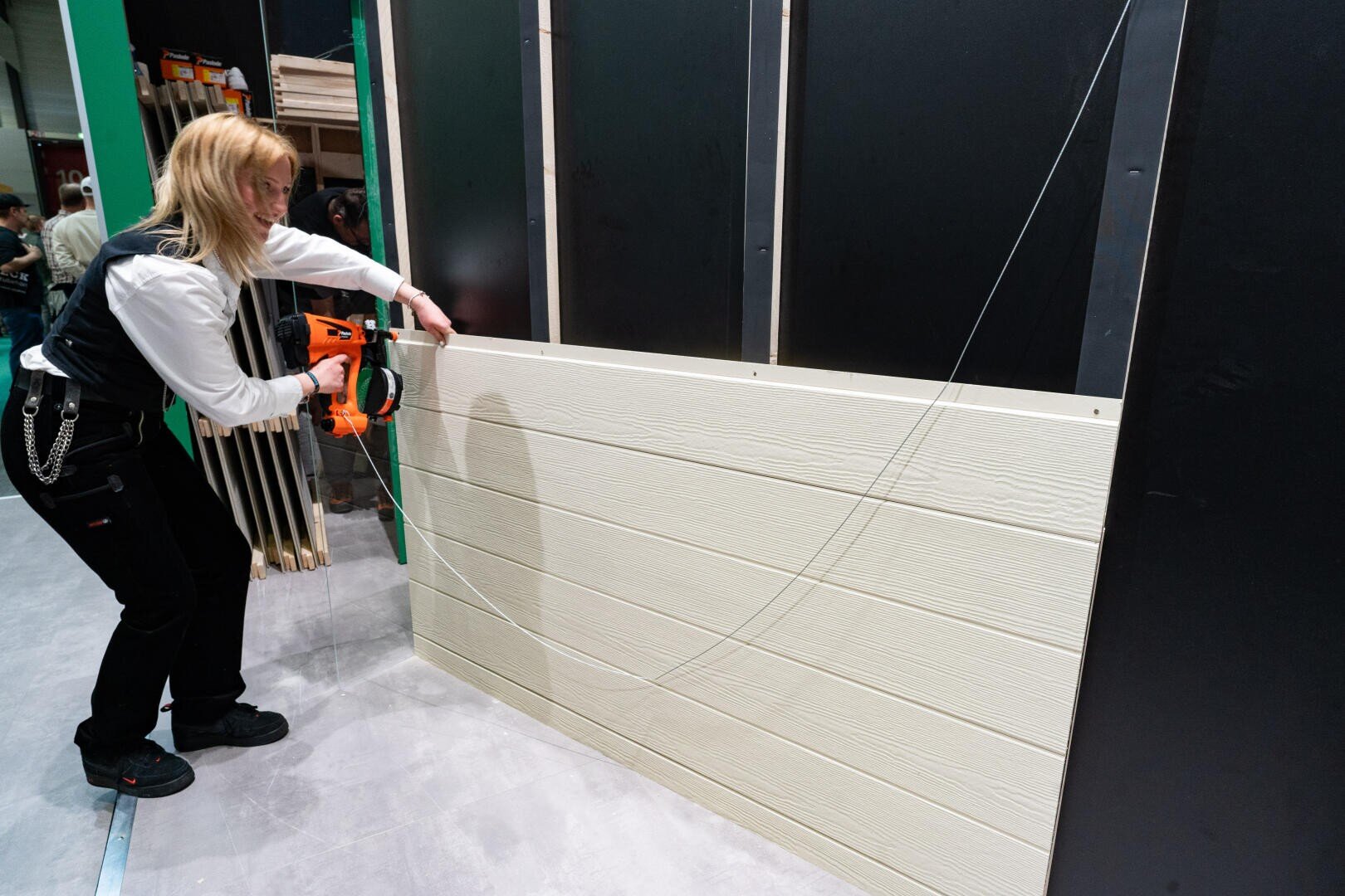 Eine Person befestigt mit einer Nagelpistole beigefarbene Holzverkleidungsplatten an einer schwarz gerahmten Wandstruktur, während im Hintergrund Werkzeuge und Baumaterialien zu sehen sind.