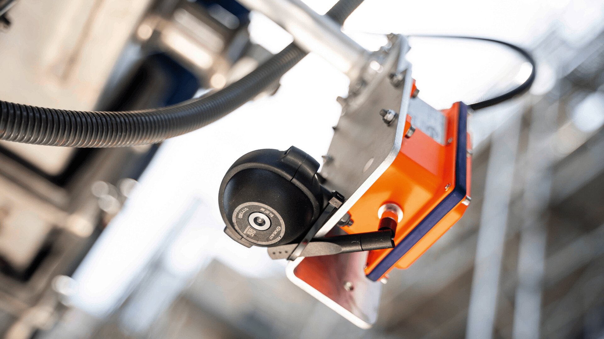 Nahaufnahme einer industriellen Sicherheitskamera, die an einer orangefarbenen Metallhalterung montiert ist, mit angeschlossenen Kabeln und einem unscharfen Hintergrund aus Strukturelementen.