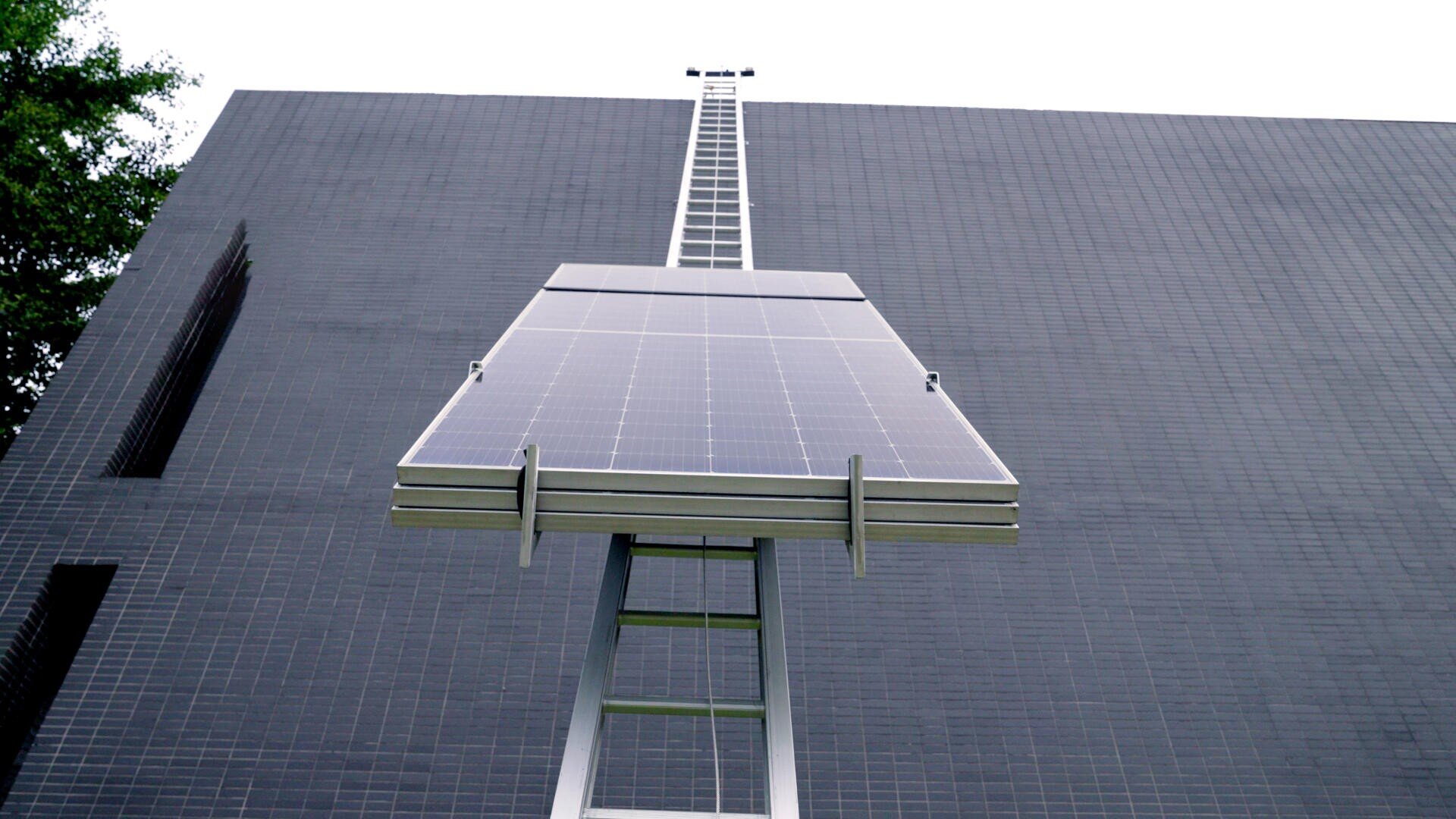 A large solar panel mounted on a metal ladder structure is positioned against the side of a dark-tiled building, with green tree foliage visible on the left edge of the image.