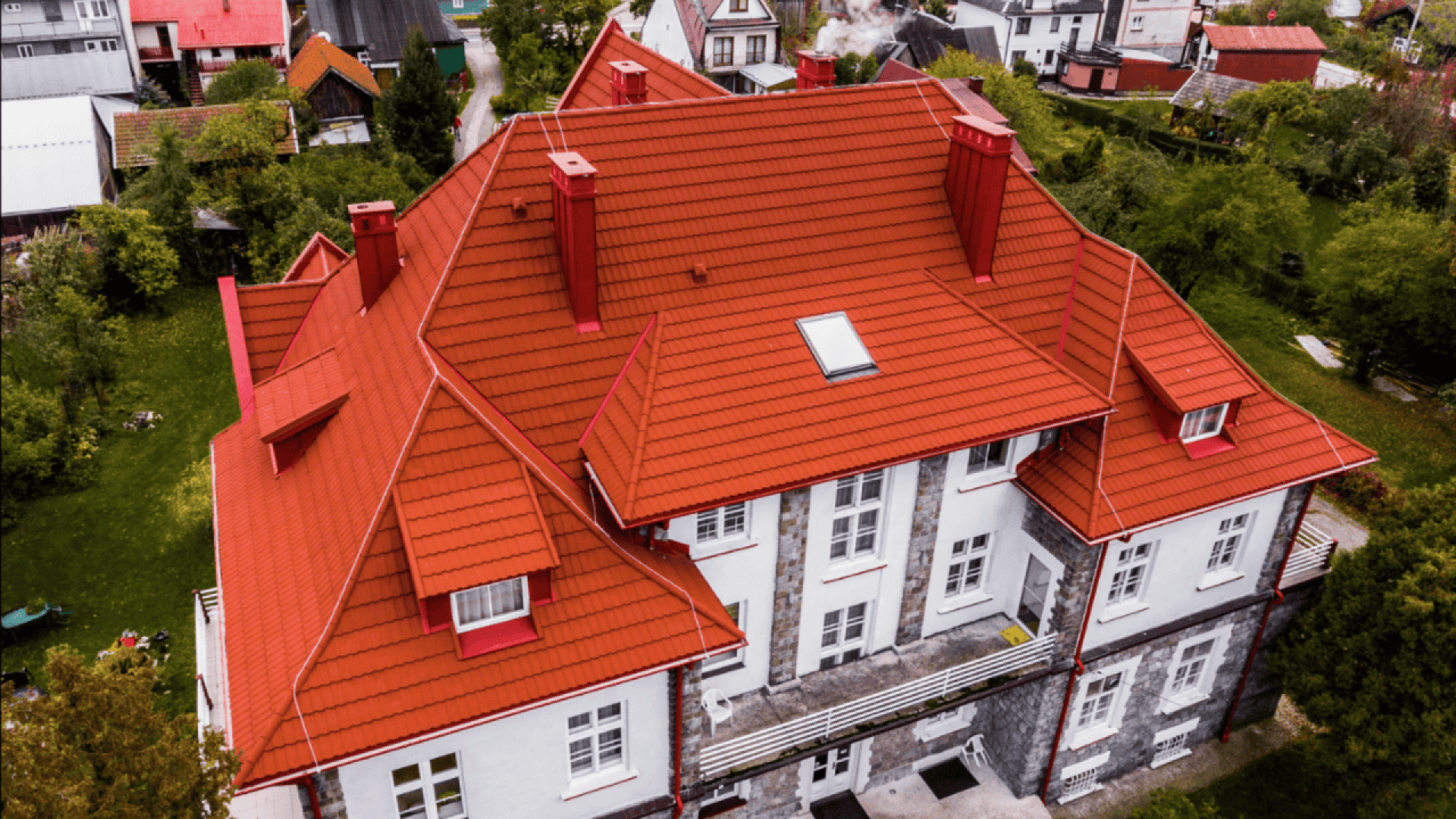 Aerial view of a large white house with stone accents and a bright red gabled roof, surrounded by green trees and other homes in a neighborhood.