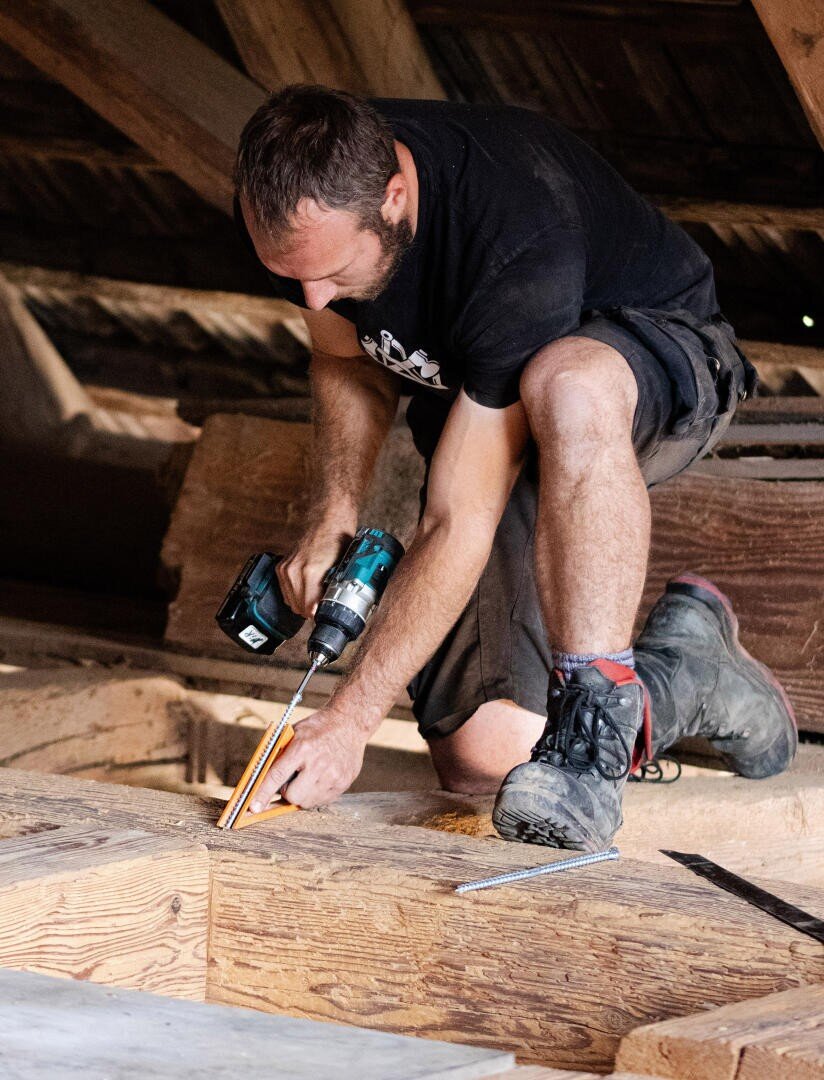 Ein Mann kniet auf den Holzbalken eines Dachbodens, hält in der einen Hand einen Akkubohrer und benutzt mit der anderen Hand ein Maßband, um das Holz zu vermessen, und arbeitet konzentriert an einem Bau- oder Renovierungsprojekt.