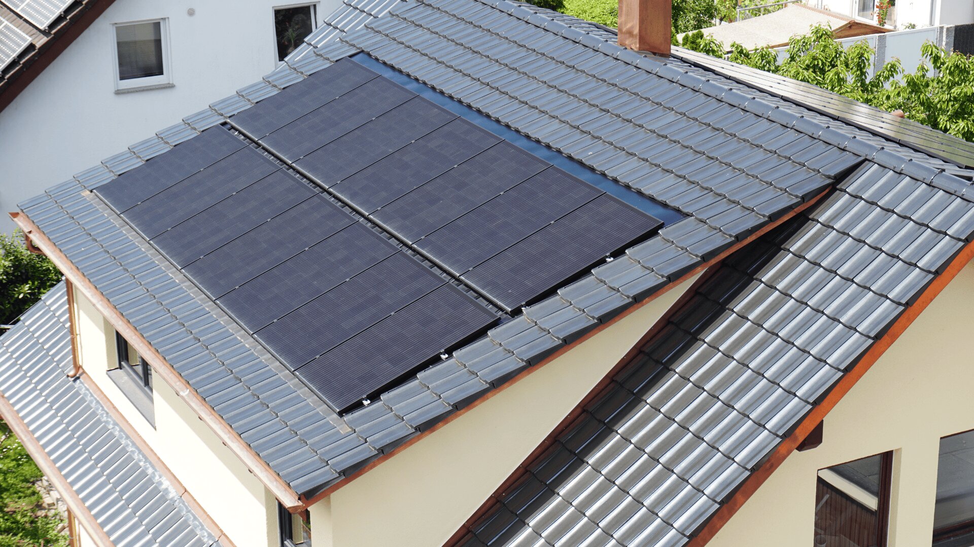 Ein modernes Haus mit einem Schrägdach, das mit mehreren Sonnenkollektoren ausgestattet ist, umgeben von Grünflächen und benachbarten Gebäuden.