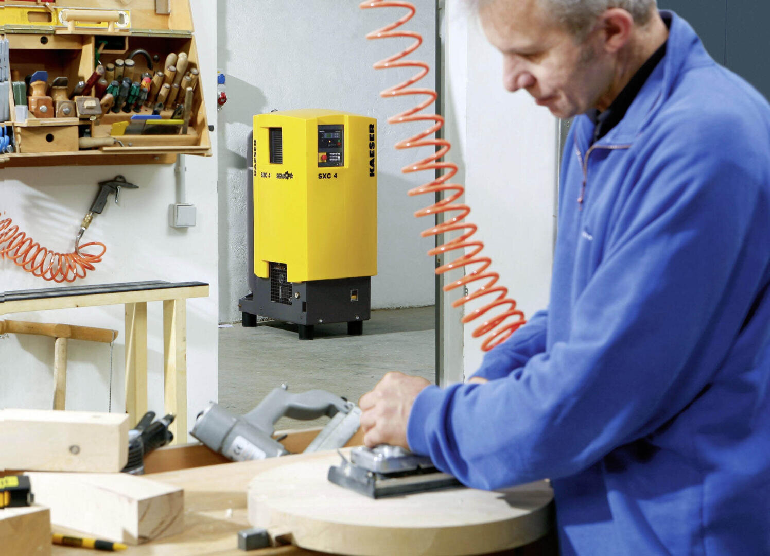 A person in a blue jacket is sanding a round piece of wood in a workshop, with woodworking tools on the wall and a yellow air compressor in the background.