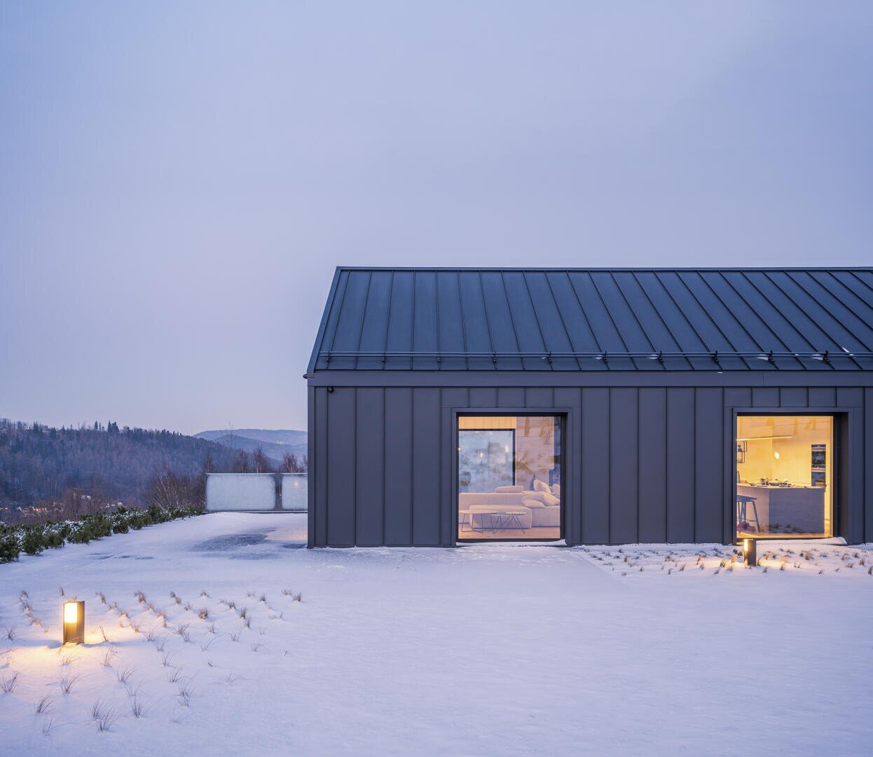 Ein modernes Haus mit einem dunklen Metalldach und zwei großen, beleuchteten Fenstern steht in einem verschneiten Hof in der Abenddämmerung. Die Außenbeleuchtung beleuchtet sanft den Schnee, und in der Ferne sind Bäume zu sehen.