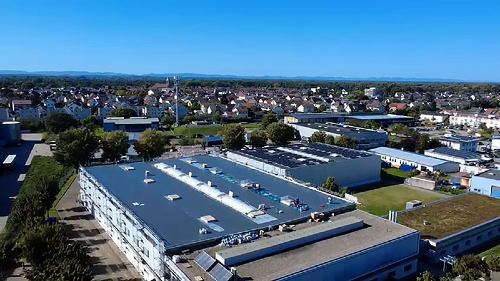 Luftaufnahme eines großen Industriegebäudes mit Flachdach, umgeben von Bäumen, anderen Gebäuden und einer Wohngegend bei klarem blauem Himmel.