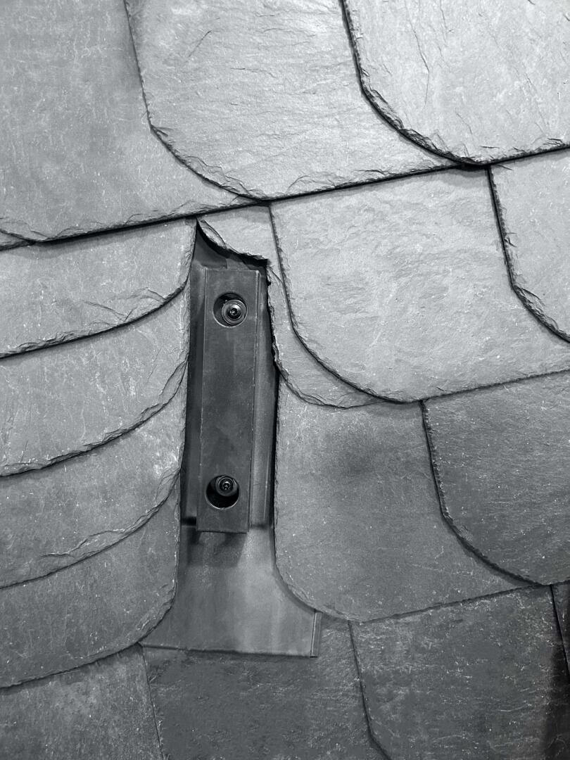 Nahaufnahme von sich überlappenden grauen Schieferdachziegeln mit einem schwarzen Metallbügel oder Eindeckrahmen, der mit zwei Schrauben befestigt und teilweise von den Ziegeln verdeckt ist.