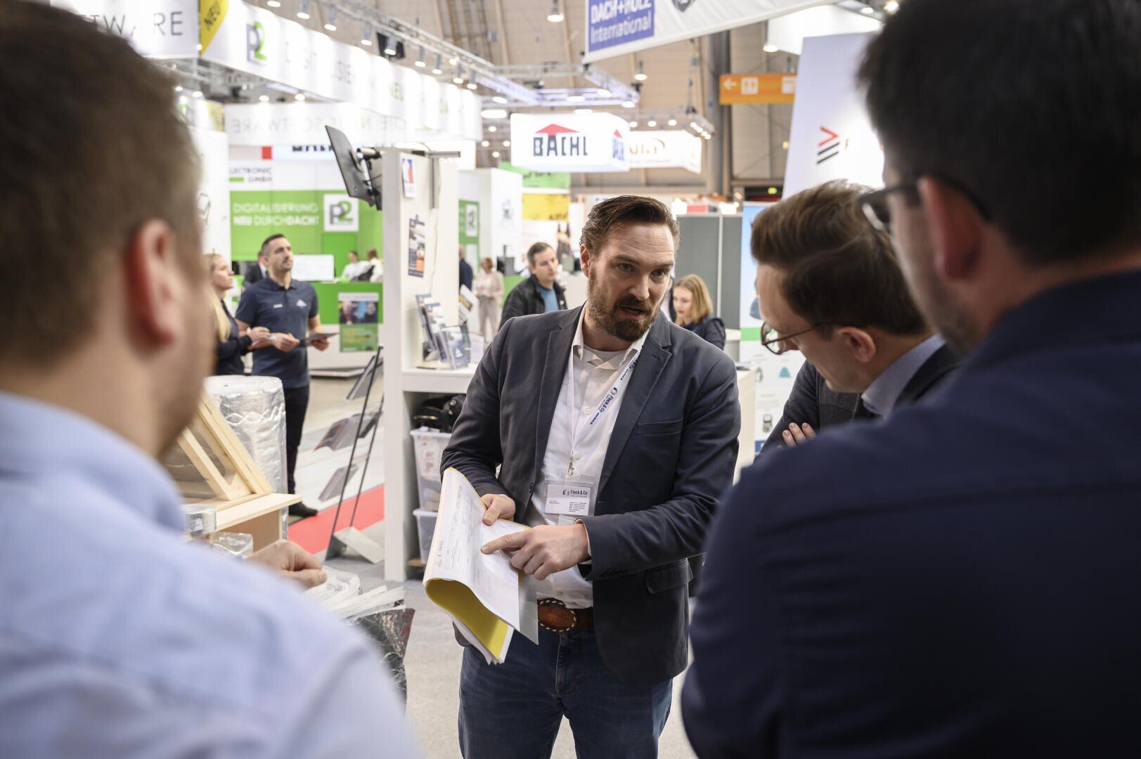 A man in business attire holds papers and talks to three men in a busy exhibition hall, with stalls, displays and people in the background.