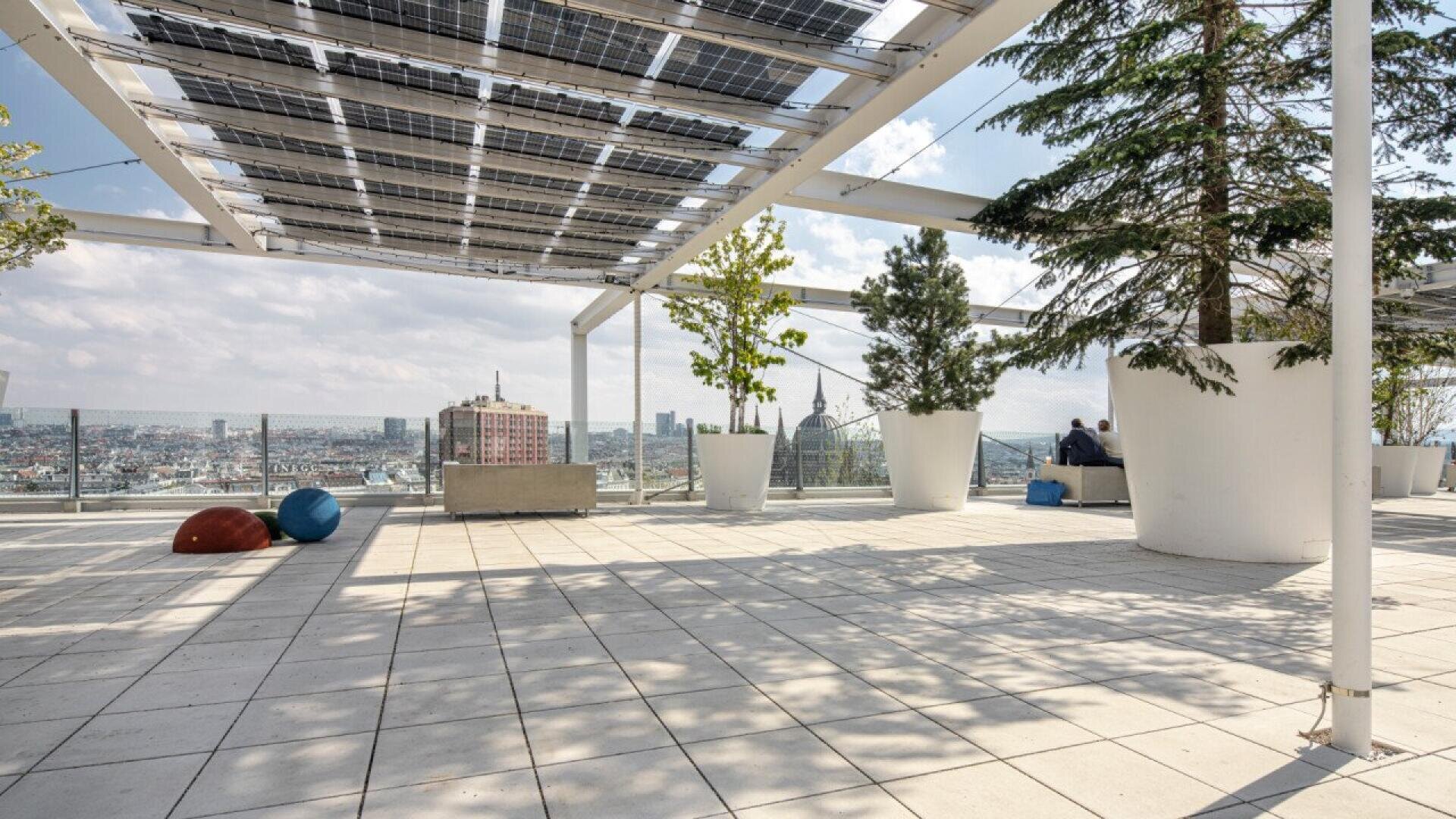 A spacious rooftop terrace with large potted trees, solar panel pergola, modern seating, and city skyline views under a partly cloudy sky.